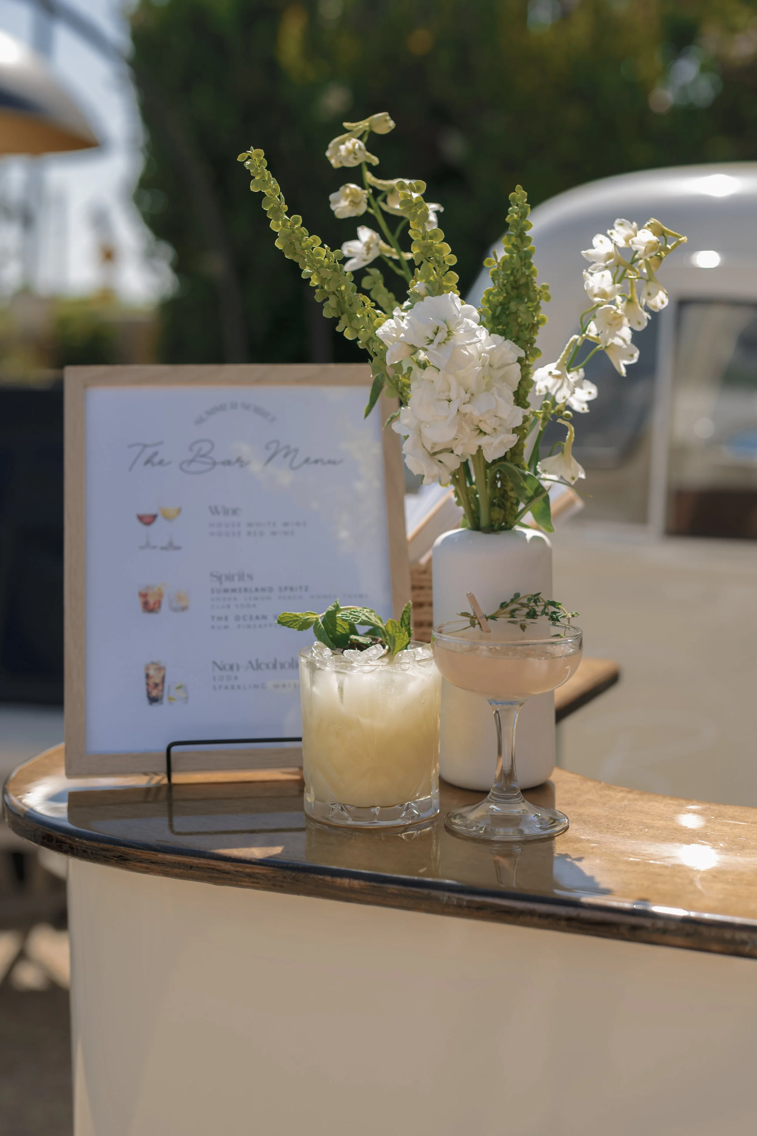 A small outdoor bar setup with a white vase containing white flowers and greenery, a cocktail in a glass, another drink in a coupe glass, and a menu sign on a wooden surface, with blurred trees and a white vehicle in the background.