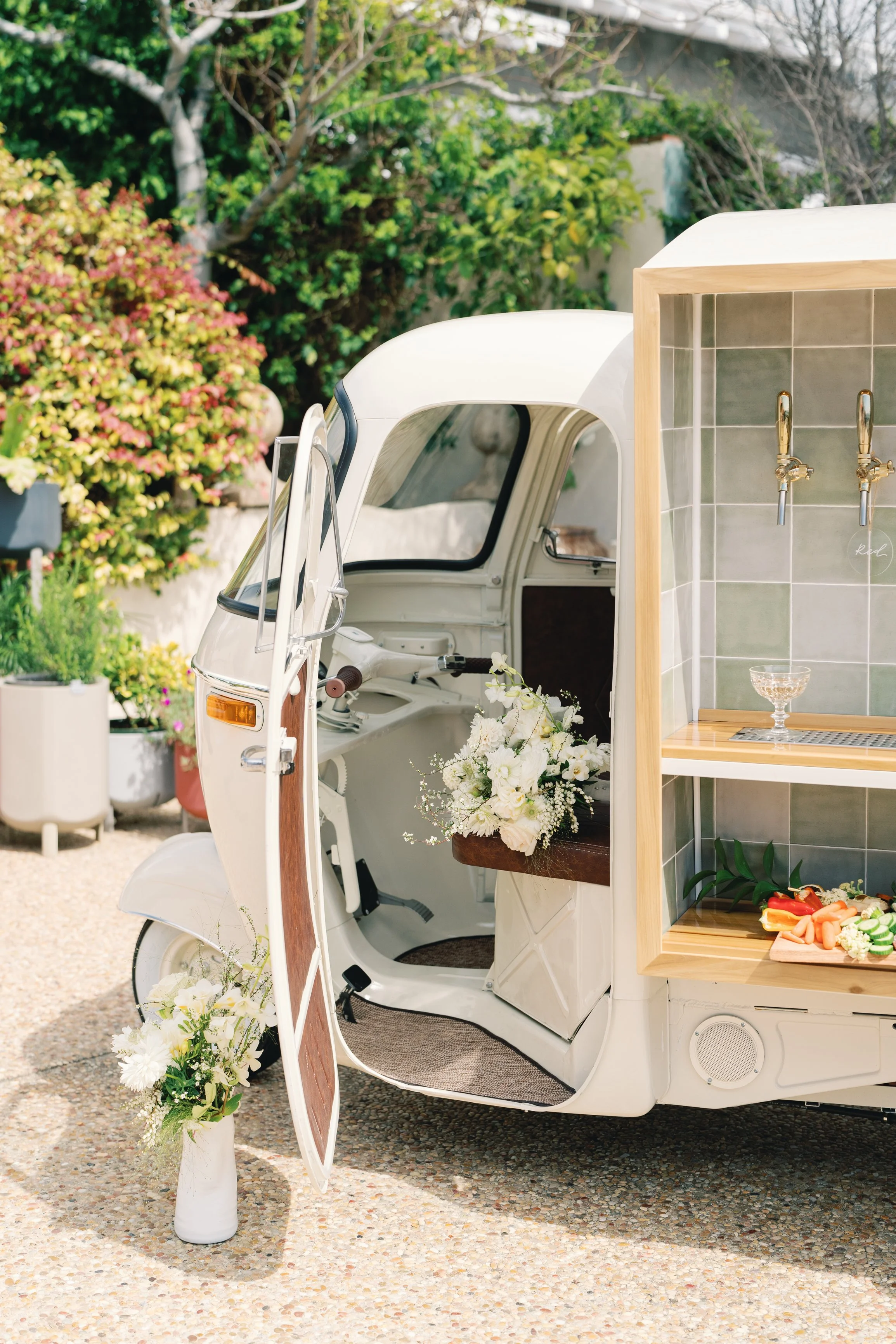 A white vintage three-wheeled vehicle converted into a food or drink stand, decorated with white flowers and greenery, with a small bar area and fresh vegetables displayed.