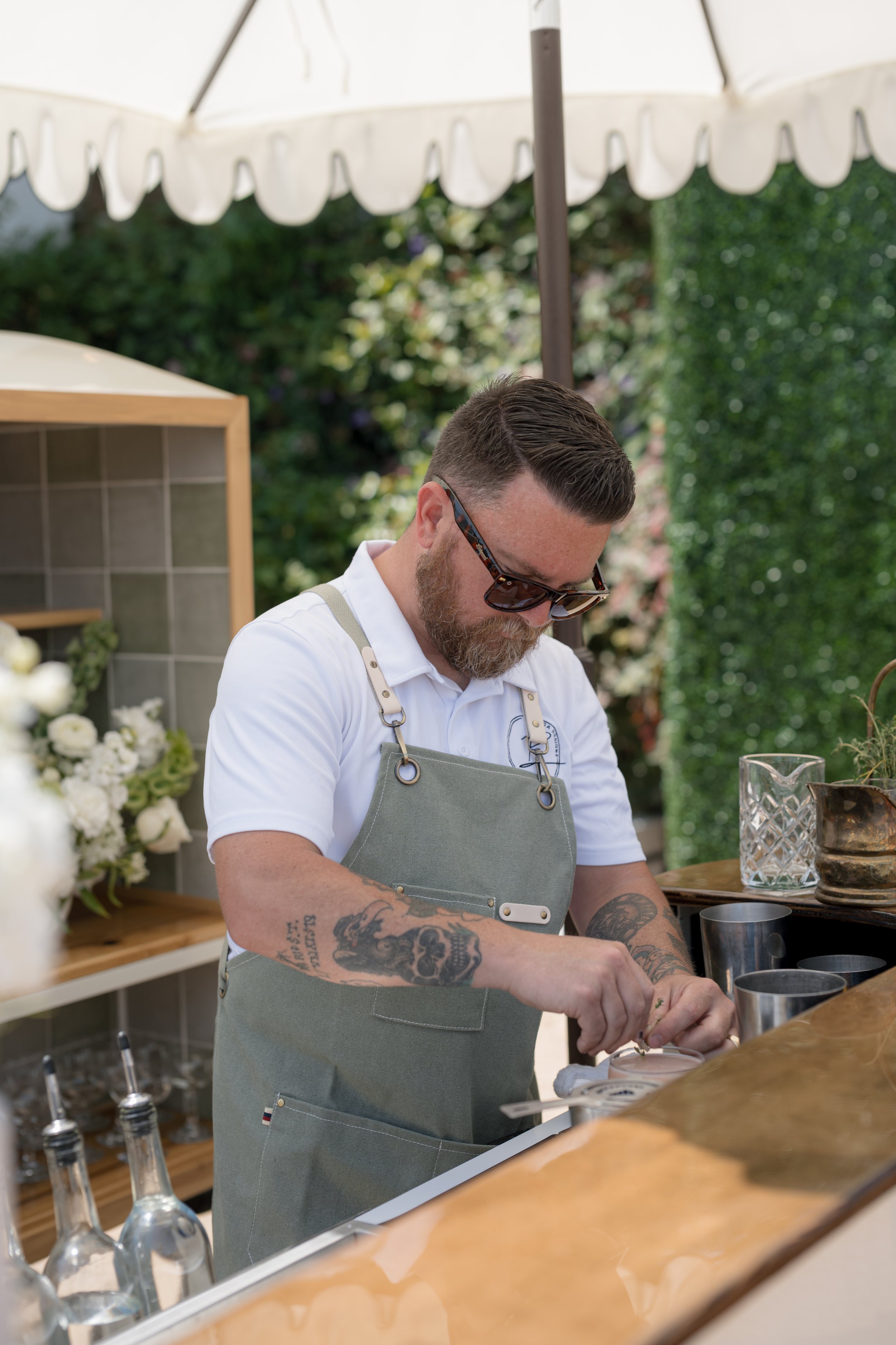 A man with tattoos and sunglasses working behind a flower and beverage stand at an outdoor event.