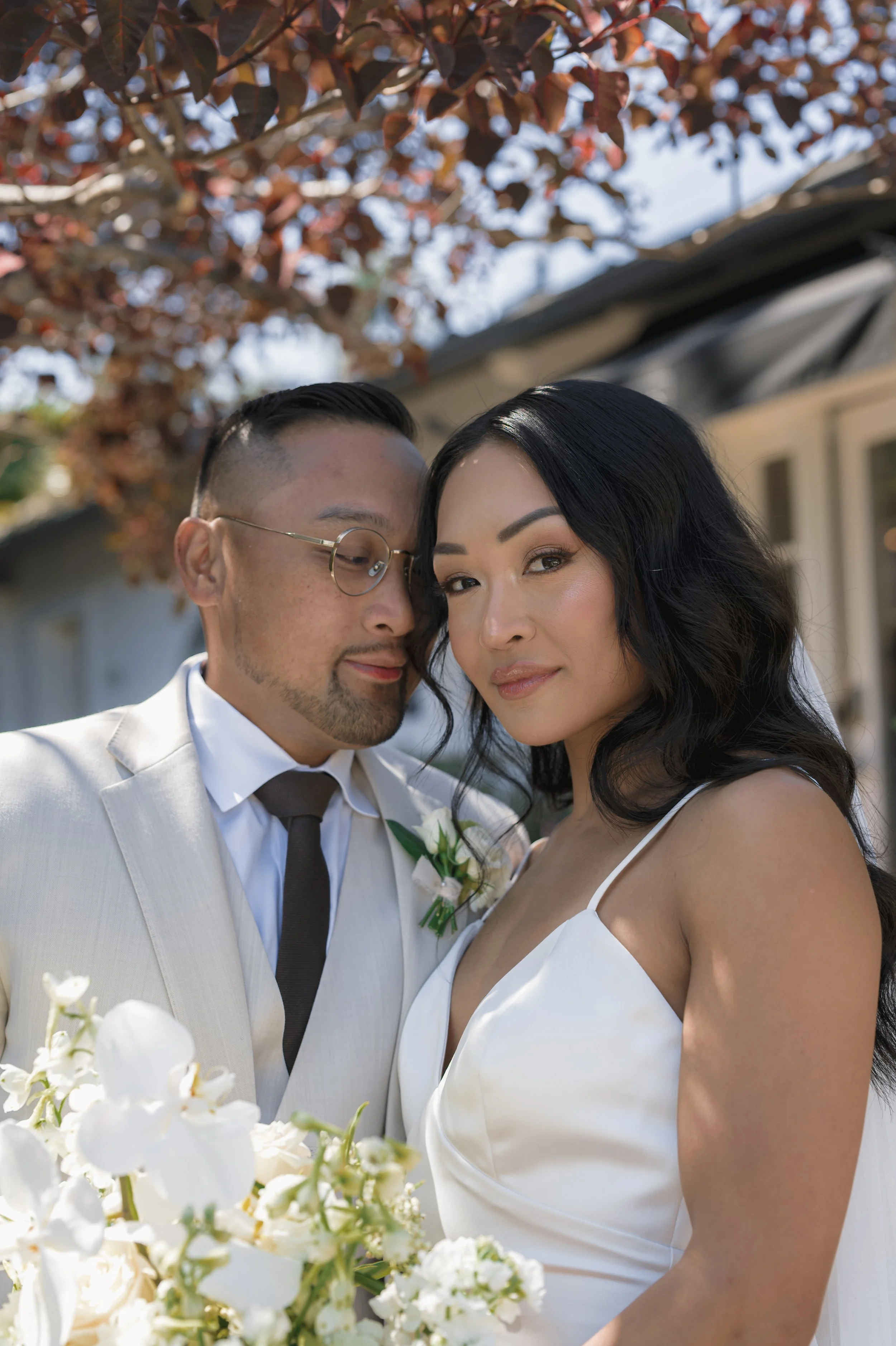 A newlywed couple standing outdoors under a tree with red leaves. The groom is wearing glasses, a light-colored suit, and a dark tie, with a white boutonniere. The bride is wearing a white dress with spaghetti straps, and holding a bouquet of white f