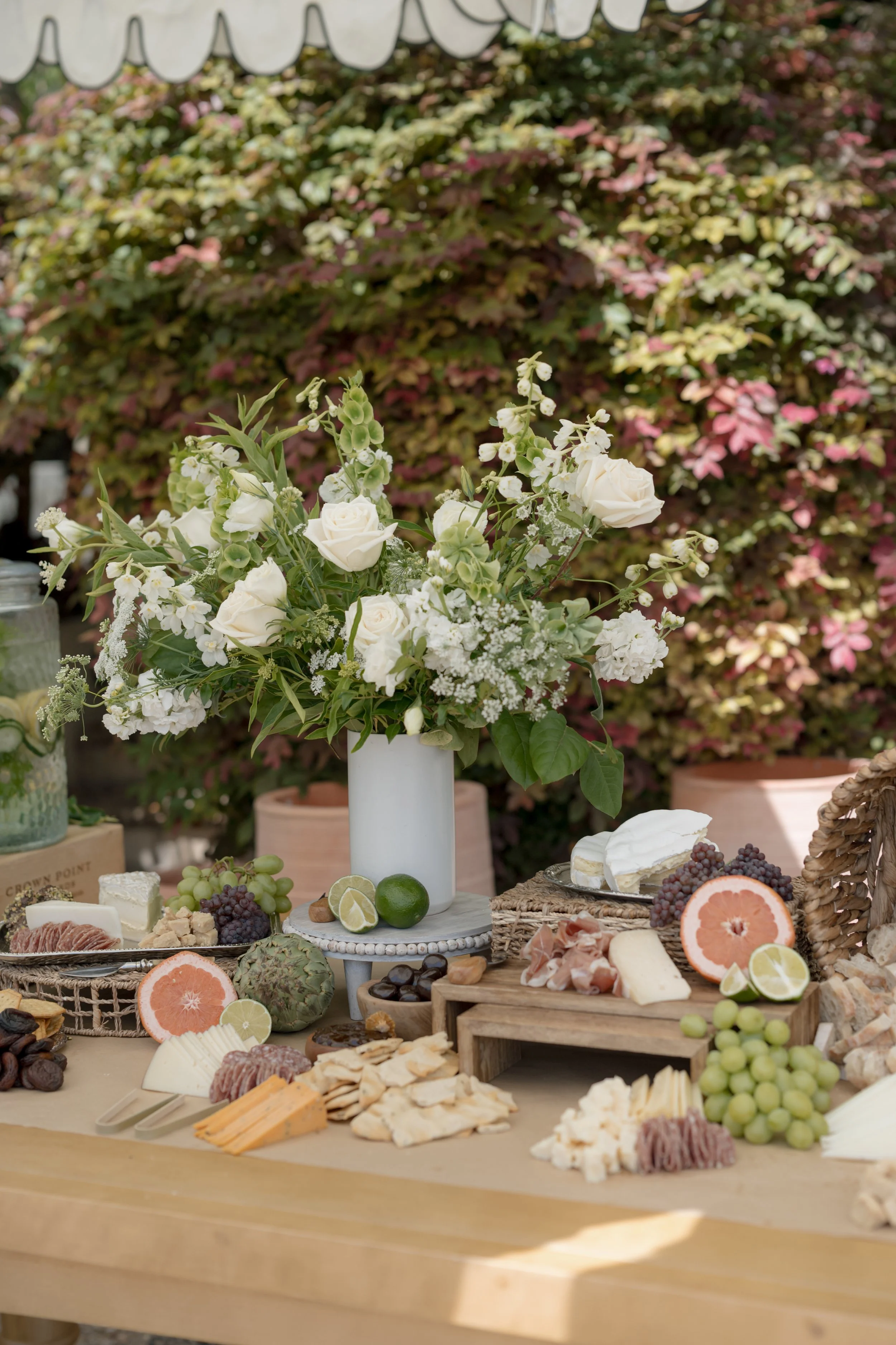 A cheese and fruit platter with a floral centerpiece on a wooden table outdoors.