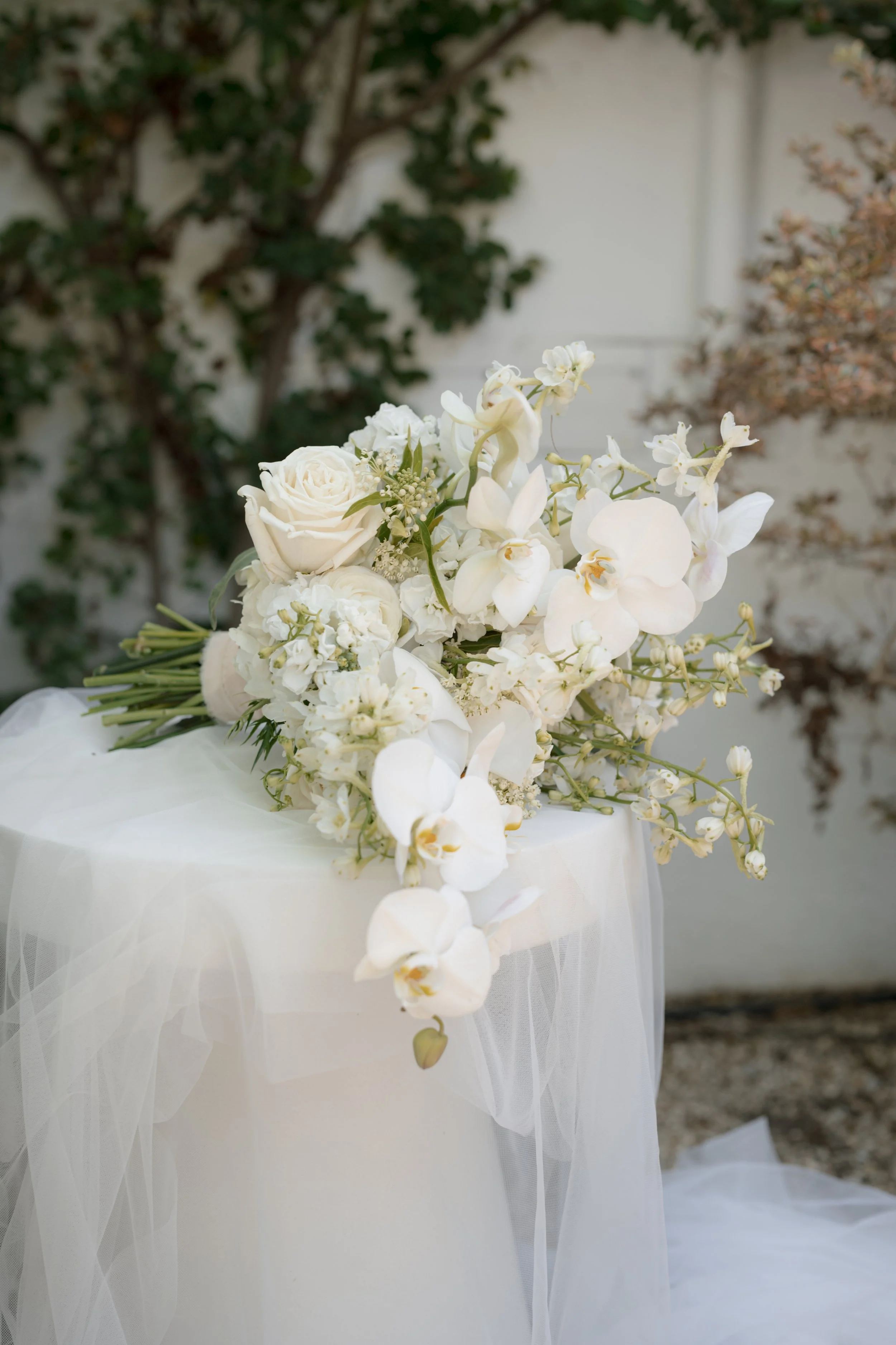 White floral bouquet with roses, orchids, and other flowers on a table draped with white fabric, outdoors with greenery in the background.