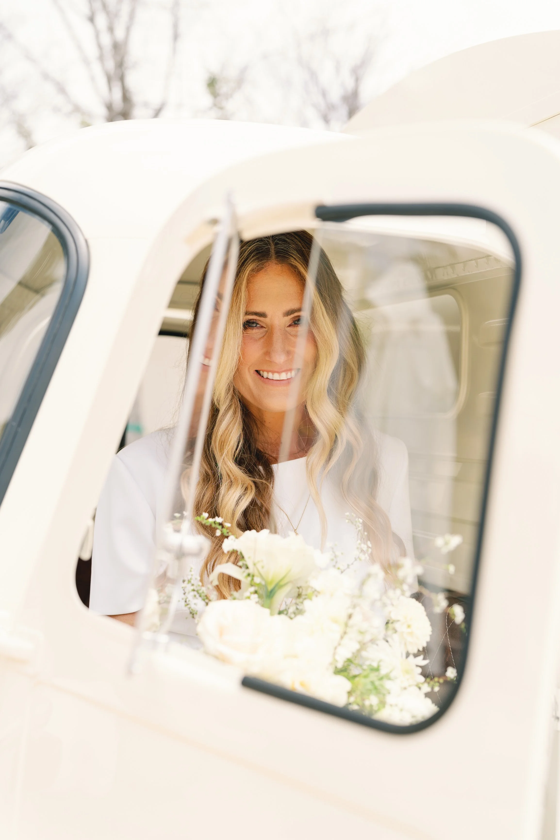 A woman with long blonde wavy hair smiling while sitting inside a vintage white truck, holding a bouquet of white flowers, with trees visible in the background.
