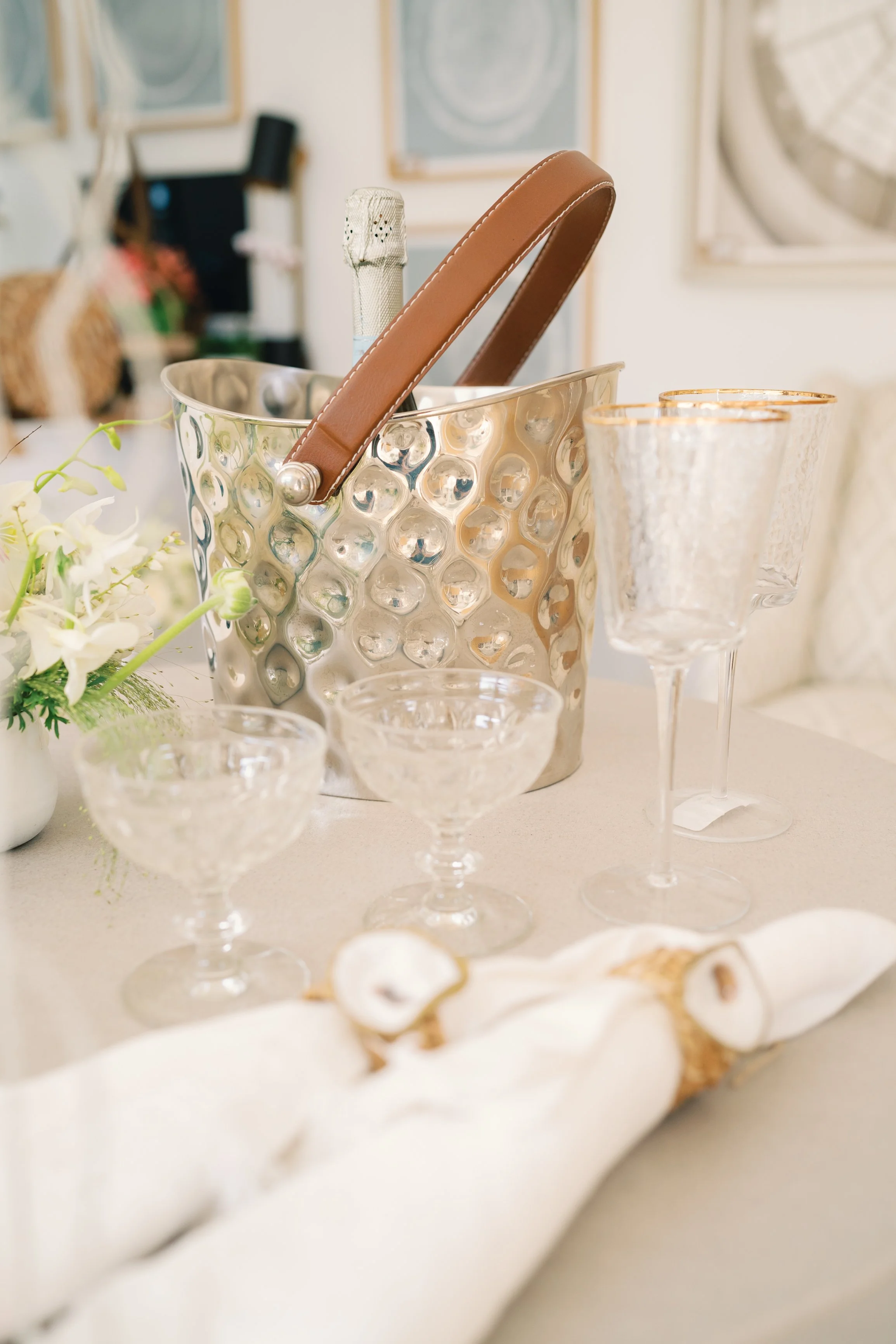 A table setting with a silver ice bucket, champagne glasses with gold rims, a white flower arrangement, and a rolled napkin. The background shows framed artwork and a wall clock.