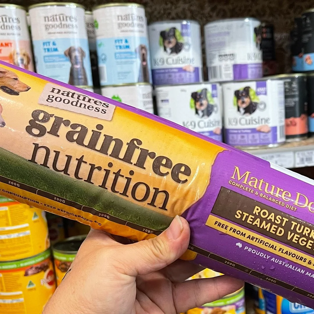 A hand holding a roll of Nature's Goodness grain-free dog food in front of shelves stocked with canned dog food. The label highlights "Grainfree Nutrition" and "Roast Turkey & Steamed Vegetables."