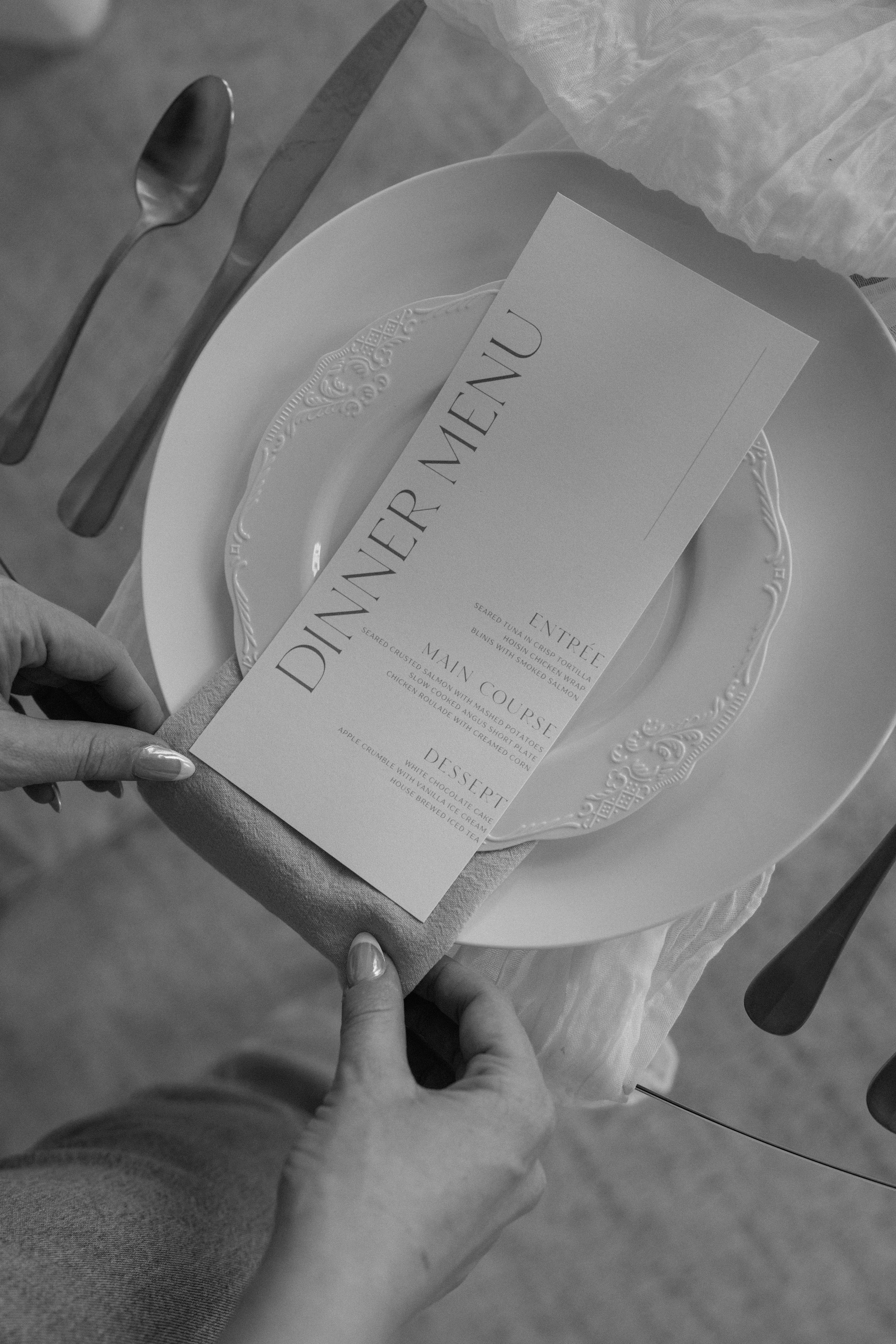 A person holding a dinner menu on a table set with a plate, cutlery, and a napkin at a restaurant.