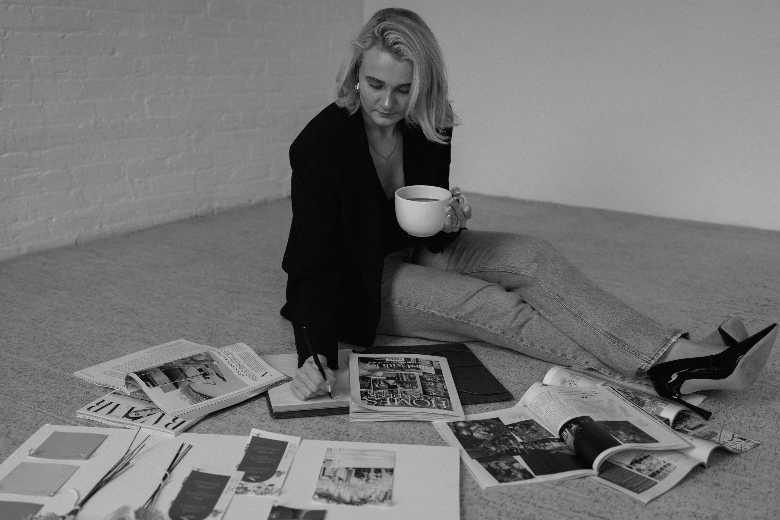 A woman sitting on the floor, writing in a notebook, surrounded by magazines, papers, and a coffee mug.