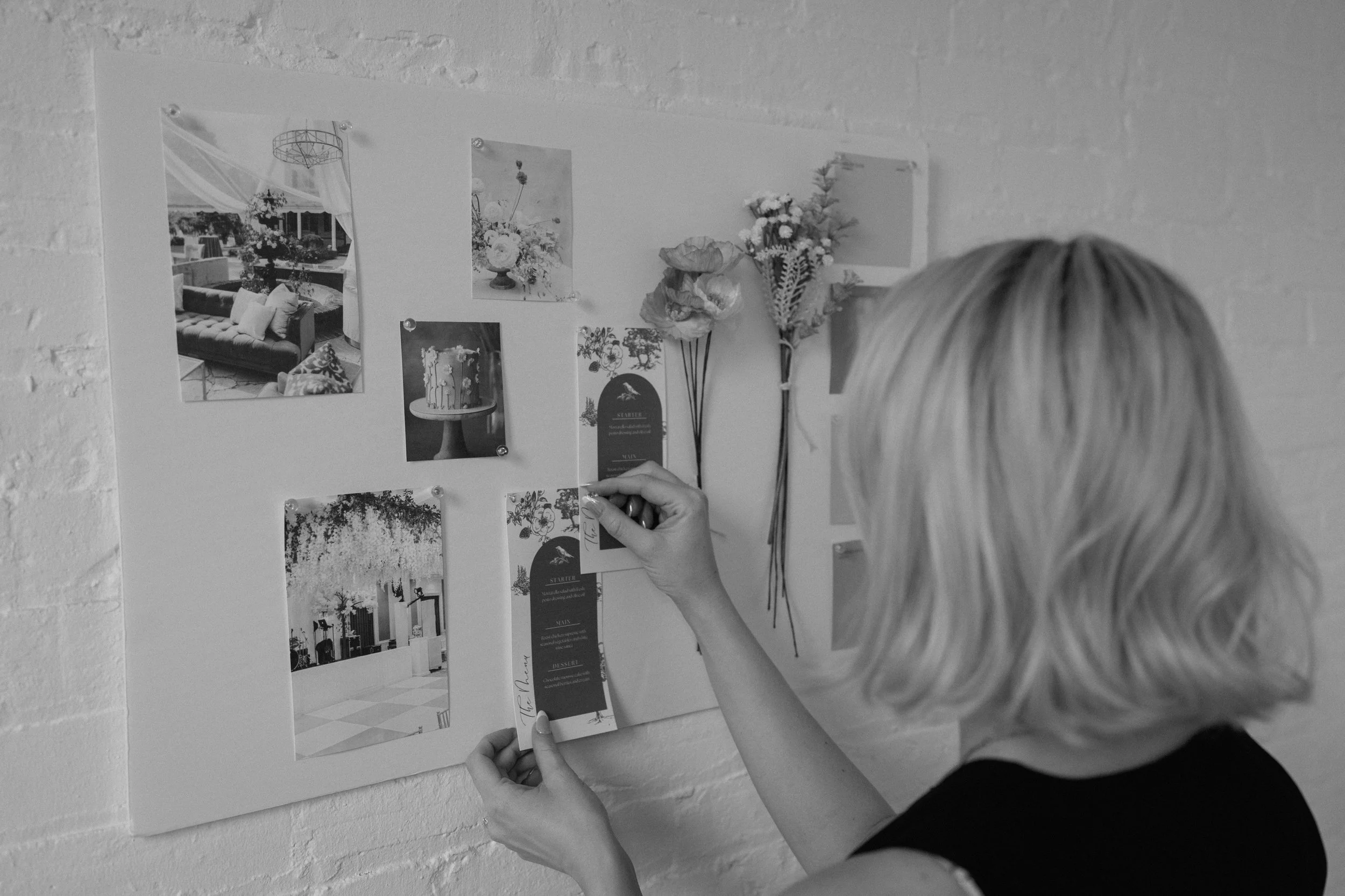A woman with short blonde hair decorating a wall with photos, flowers, and cards for an event or wedding.