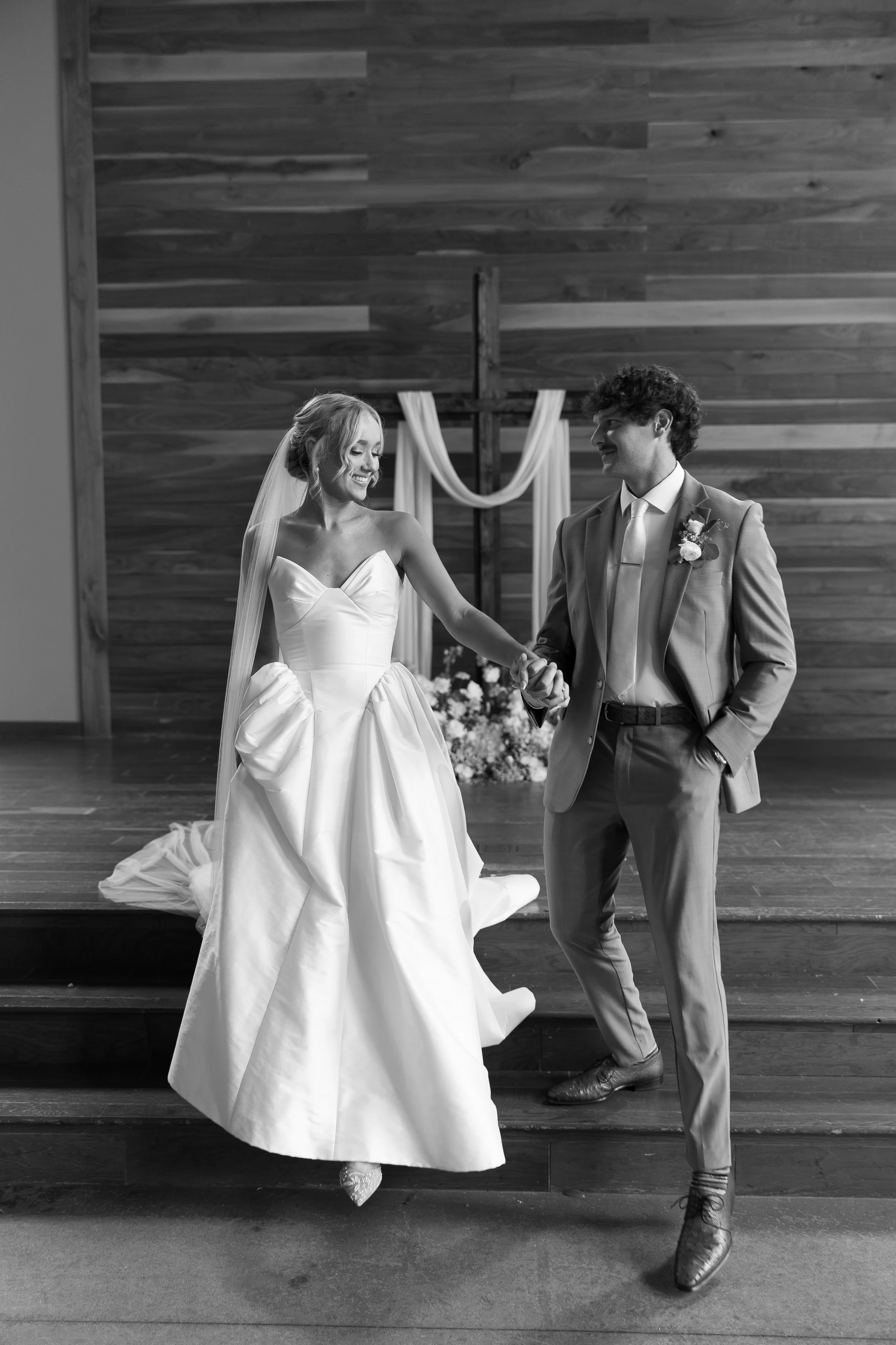A bride and groom holding hands and smiling inside a church, with a wooden cross and drapery in the background, during their wedding ceremony.
