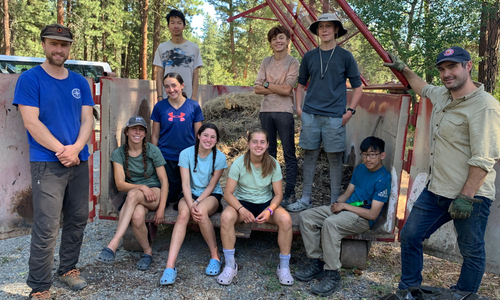 Group 8 high school kids and 2 adults standing in front of dumpster full of yard waste