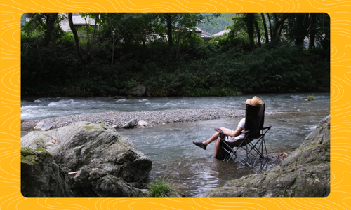 Person sitting in a camping chair in the river about ankle deep with a straw hat on