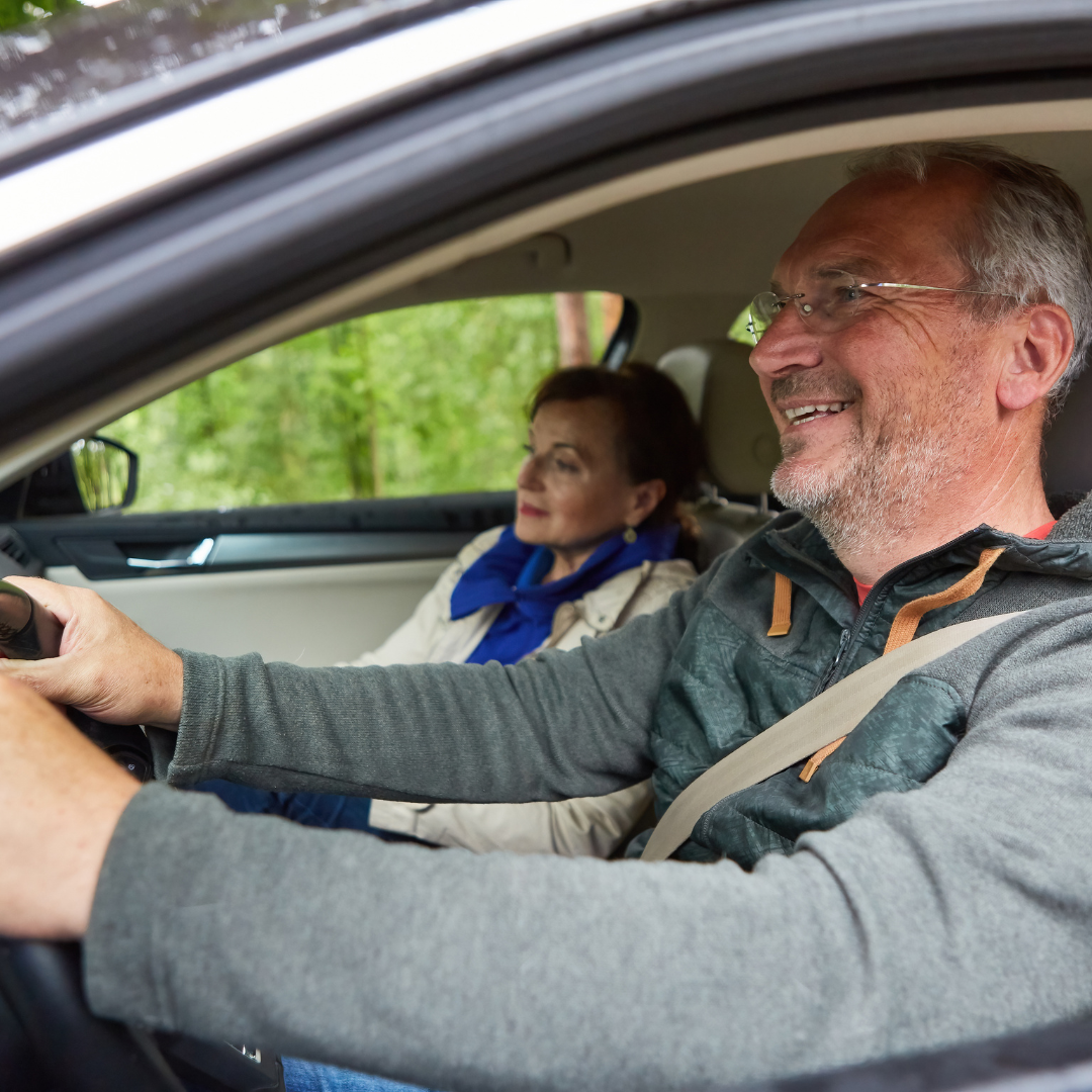 A smiling older man driving a car with a woman sitting in the passenger seat, both appearing happy, with green trees outside the car window.