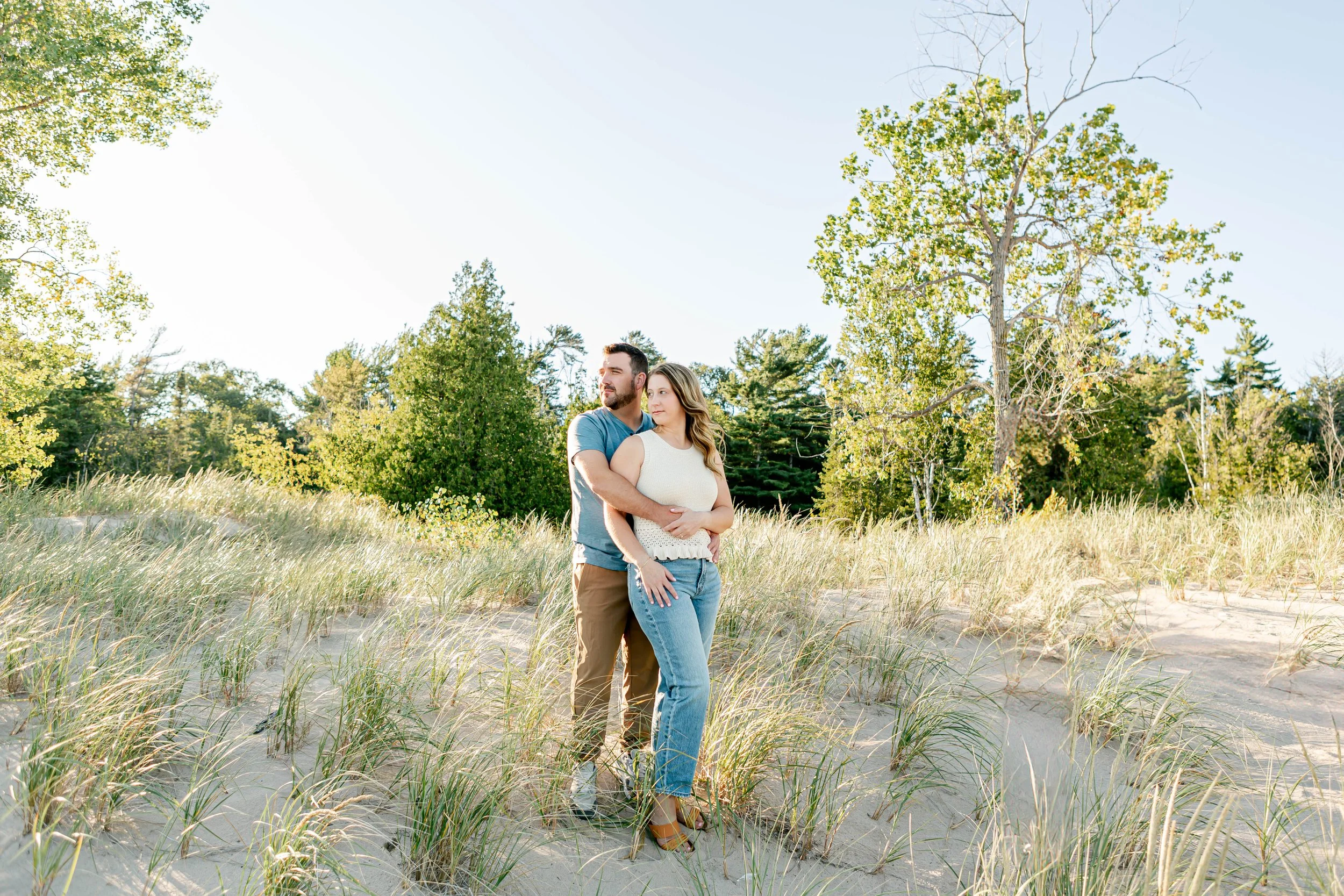 A couple standing in a sandy area with grass and trees in the background on a sunny day.