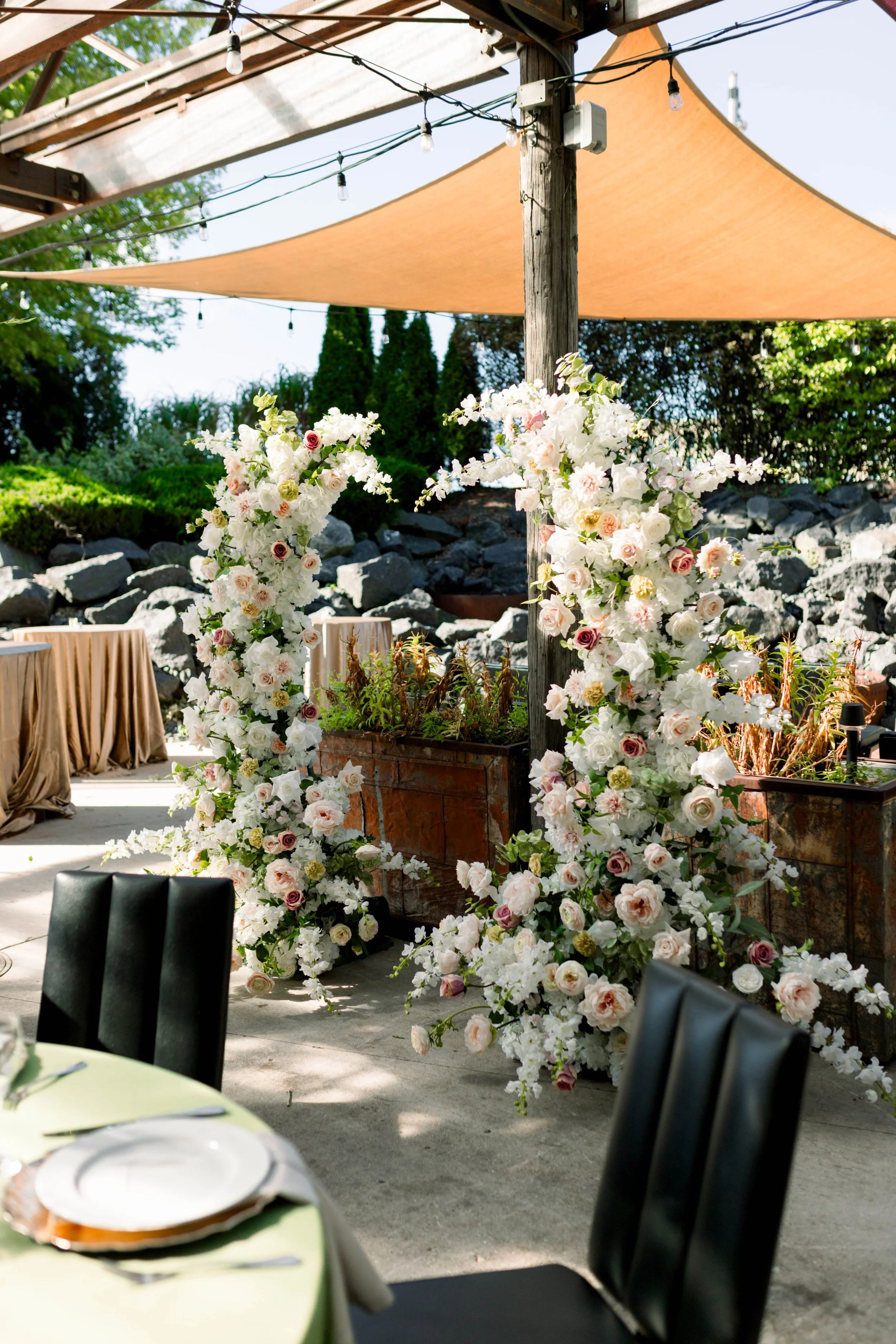 Outdoor wedding reception setup with floral arches, string lights, and tables decorated with tablecloths, under a canopy of fabric and string lights, surrounded by trees and rocks.