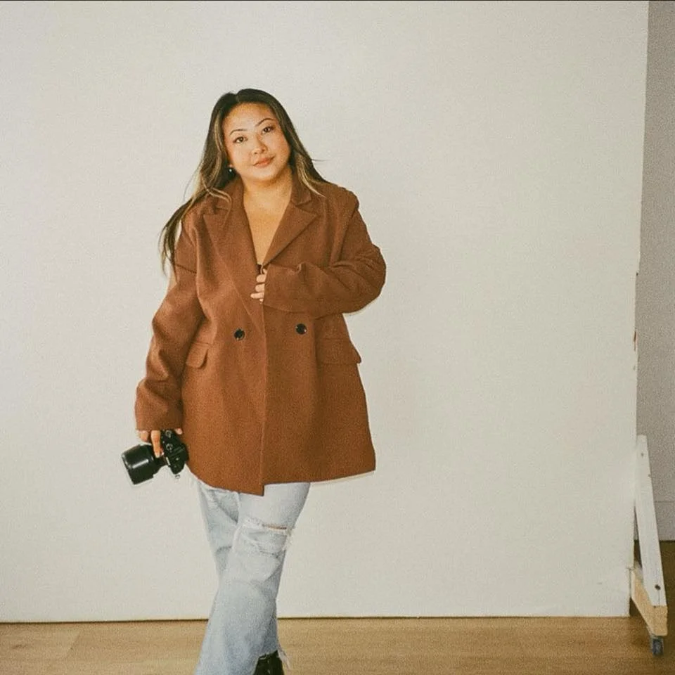 Woman in a brown blazer and ripped jeans holding a camera, standing against a plain wall.