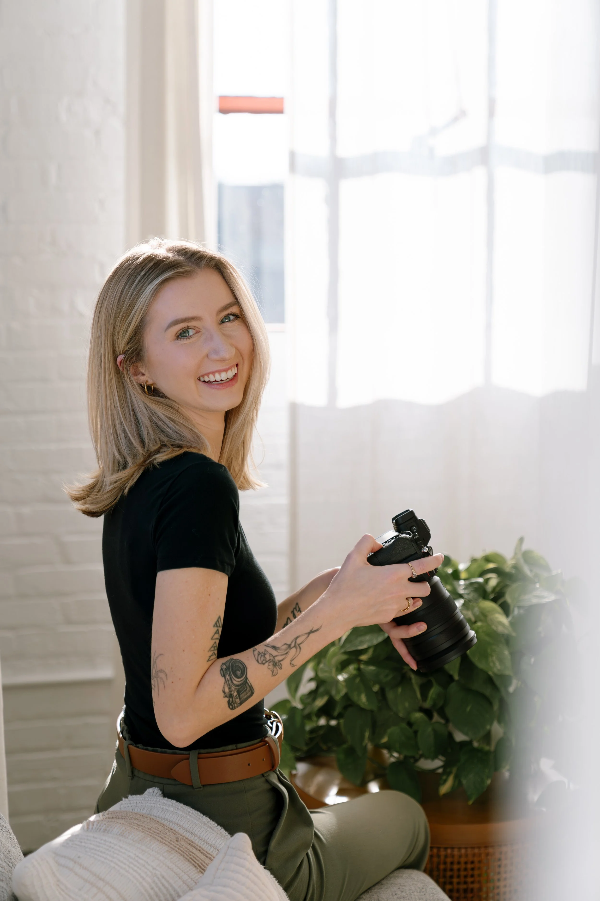 A smiling woman with blond hair, tattoos on her arms, and wearing a black shirt and green pants, sitting on a couch holding a camera in a bright room with natural light, a white brick wall, a potted plant, and white curtains.
