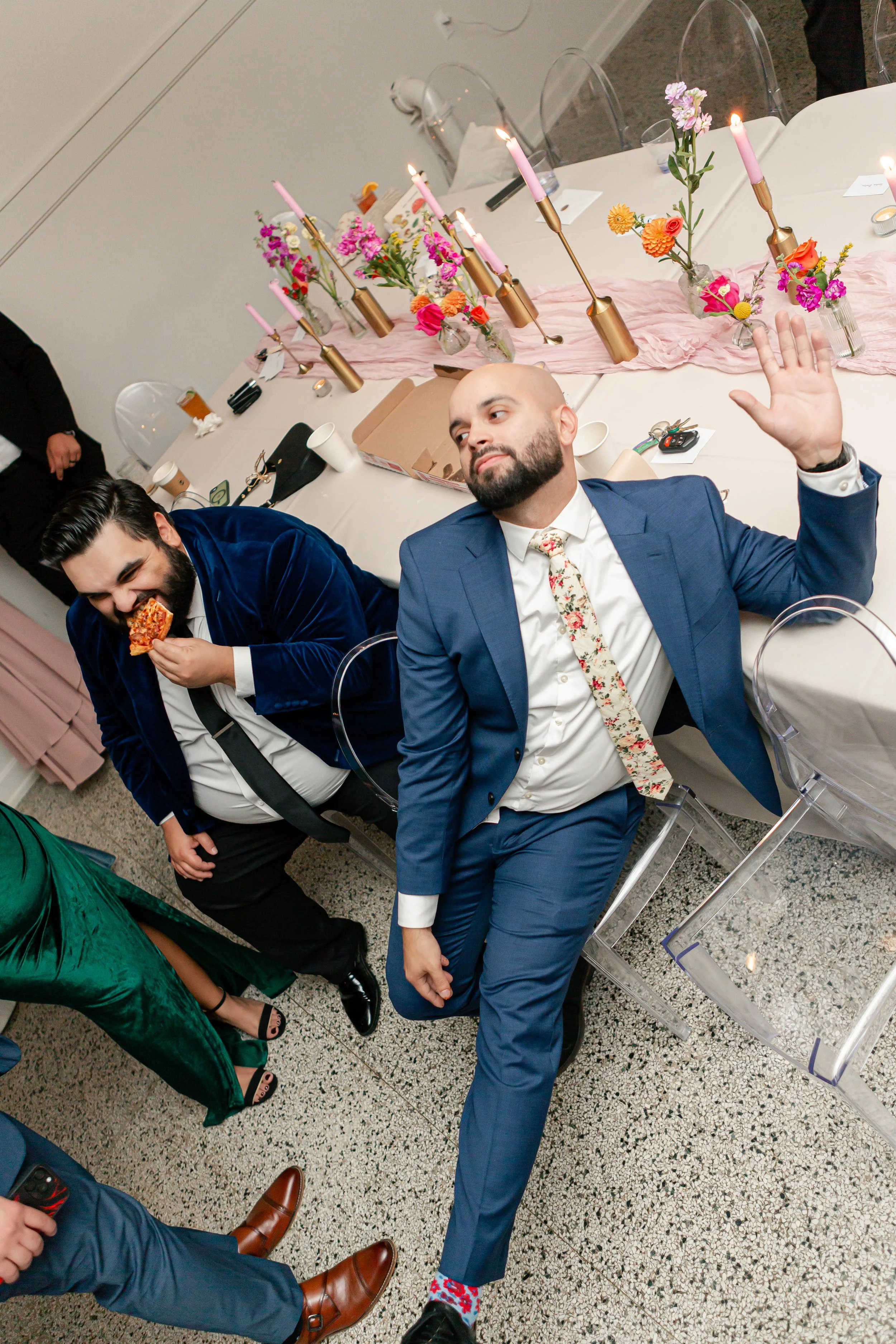 Two men in suits at a celebration dinner, one is eating pizza and the other is raising his hand.