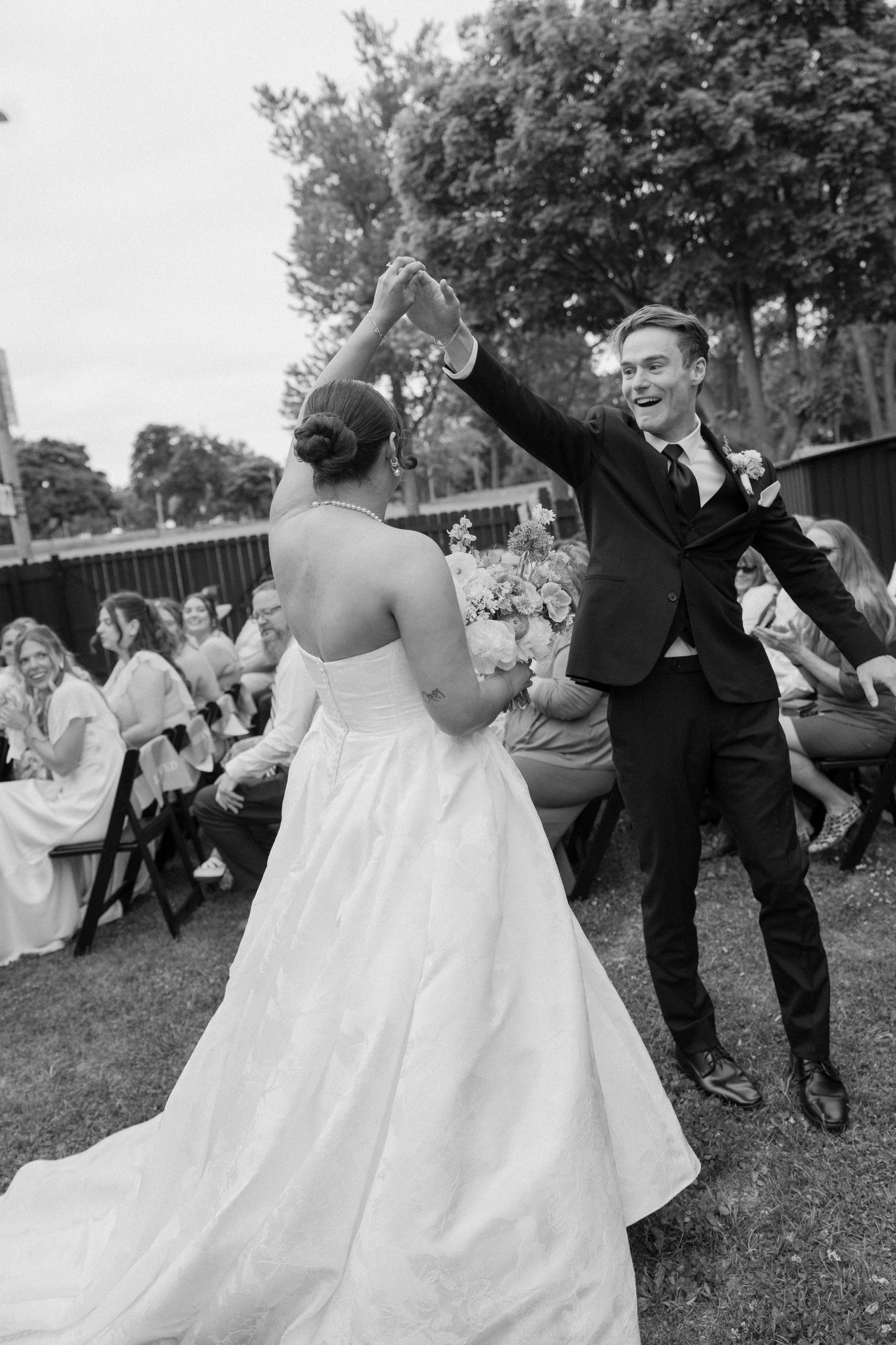 A bride and groom dancing outdoors at their wedding with guests seated nearby, trees in the background, black and white photo.