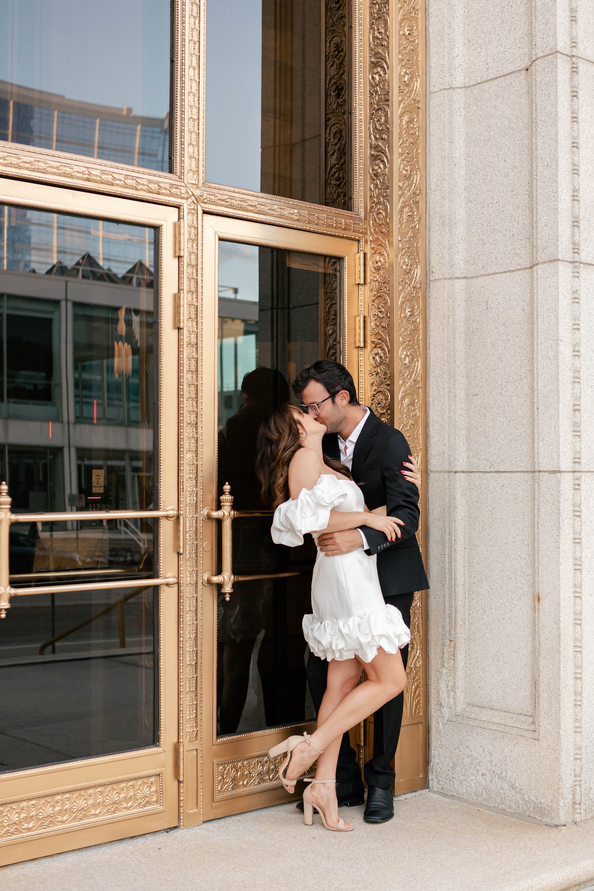 A couple kissing outside a building with ornate gold trim and large glass window