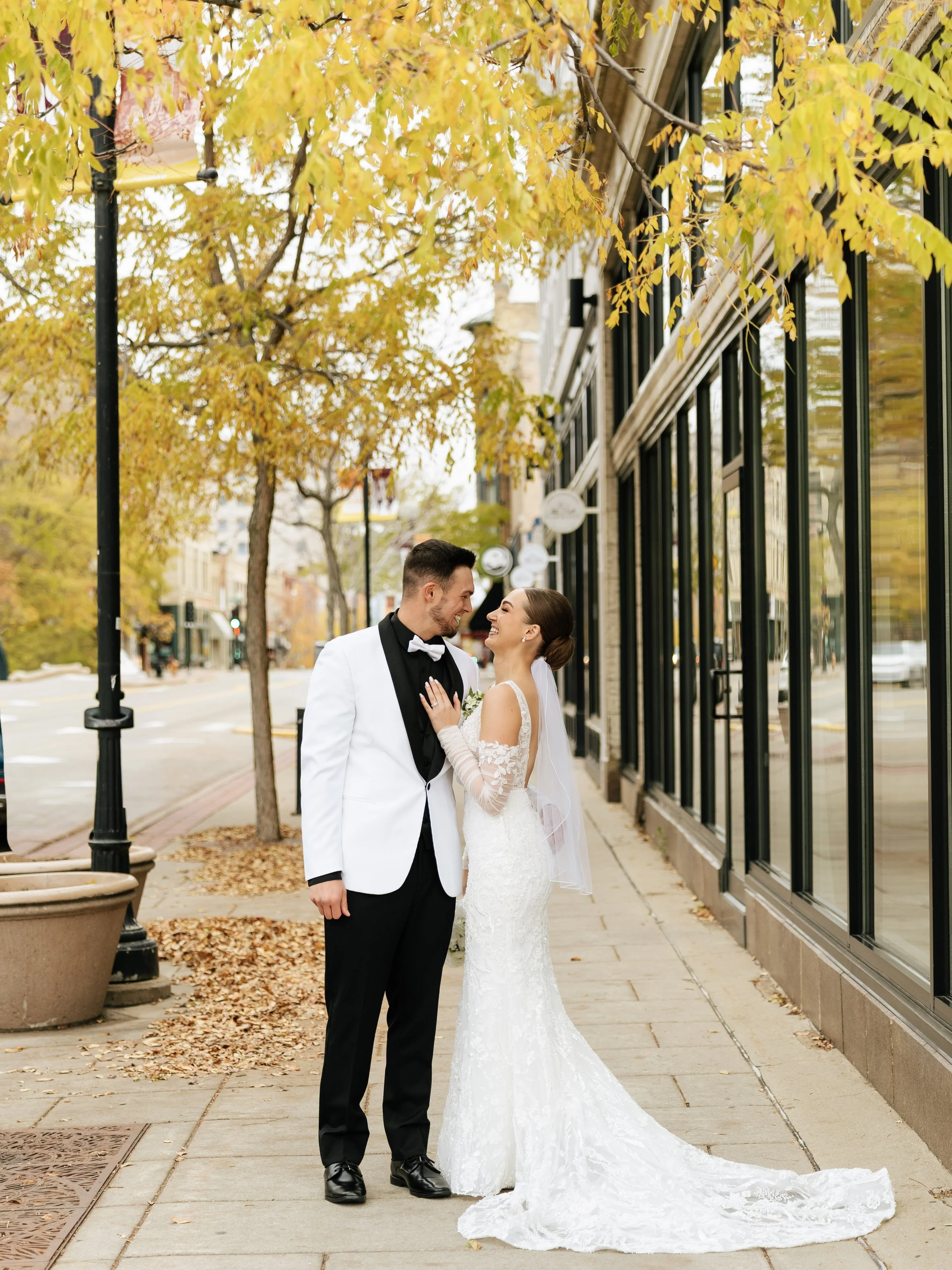A newlywed couple dressed in wedding attire standing on a city sidewalk, smiling at each other, with autumn trees and storefronts in the background.