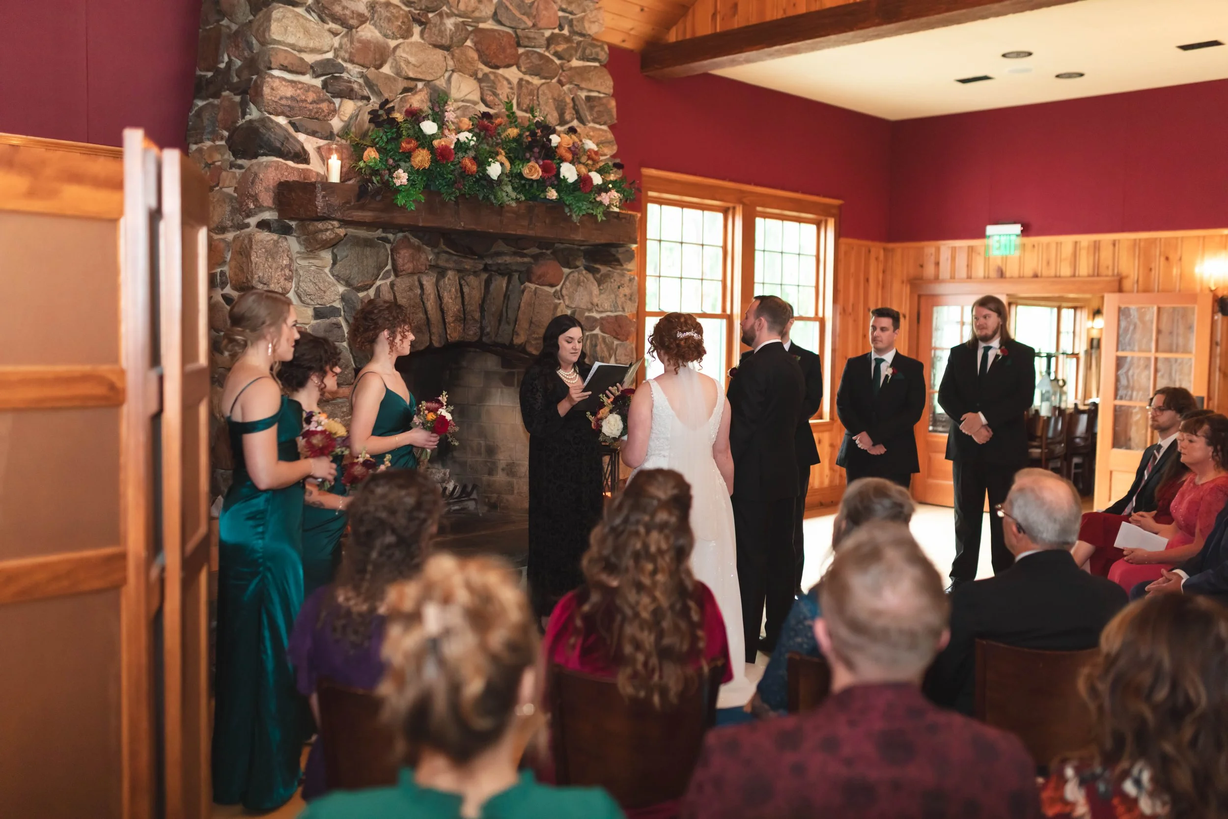 A wedding ceremony taking place inside a rustic wooden lodge with large stone fireplace, floral decorations, and large windows letting in natural light. The bride and groom are exchanging vows in front of an officiant surrounded by bridesmaids and groomsmen, with guests seated and watching.