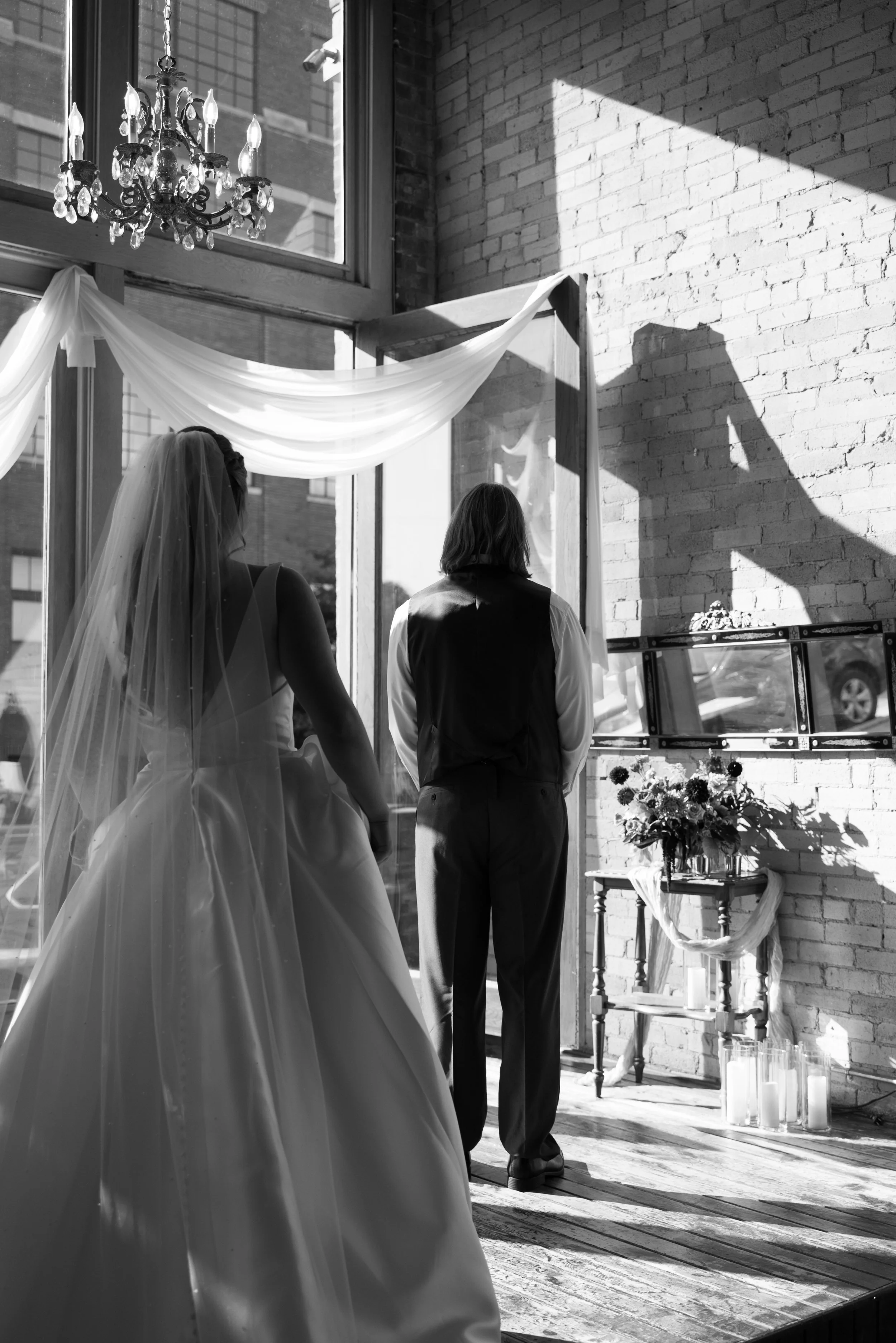 A bride and groom standing together indoors during their wedding ceremony, with sunlight streaming through large windows behind them. The bride is wearing a wedding gown and veil, and the groom is dressed in a vest and tie. The scene features a chandelier, draped fabric, floral arrangements, and candles.