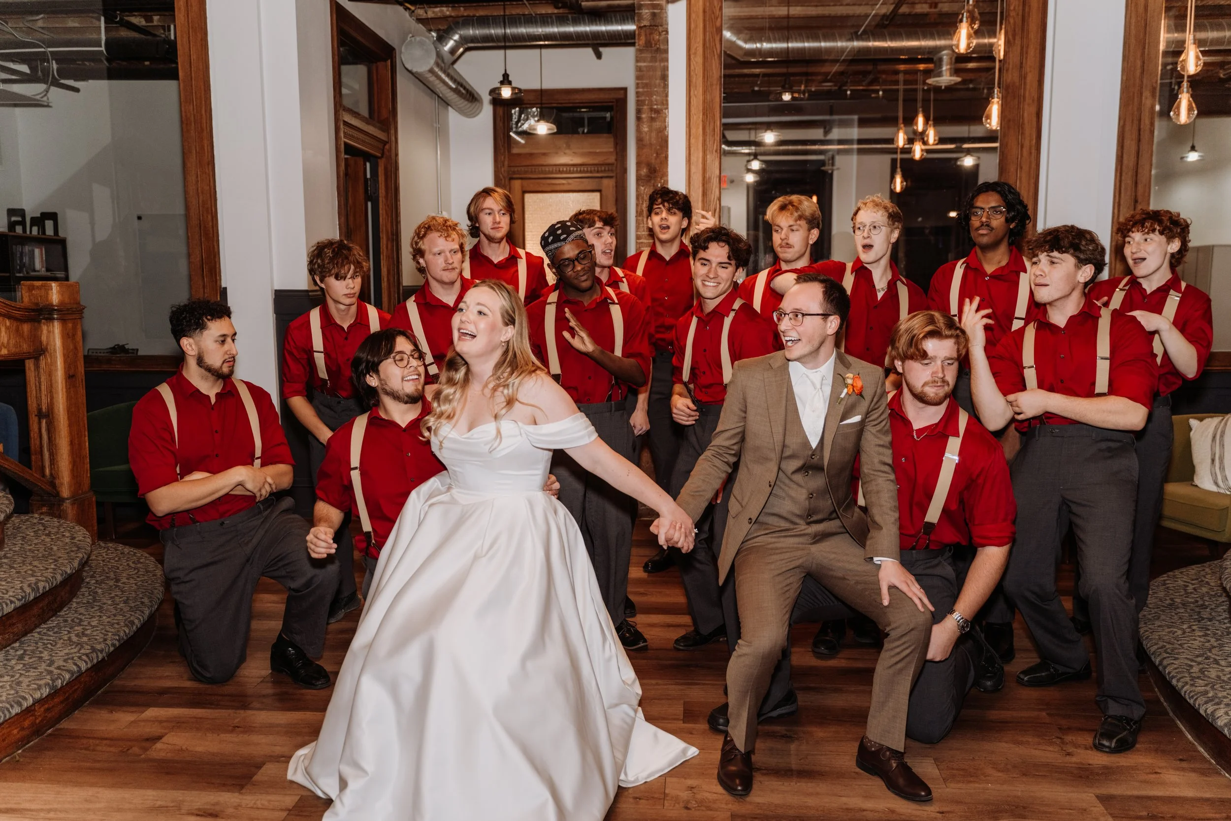 A group of young people, most wearing red shirts with beige suspenders, gathered in a warmly lit room with wooden accents and exposed ductwork. A smiling woman in a white dress is sitting in the front, holding hands with a man in a beige suit, while others are standing and engaging.