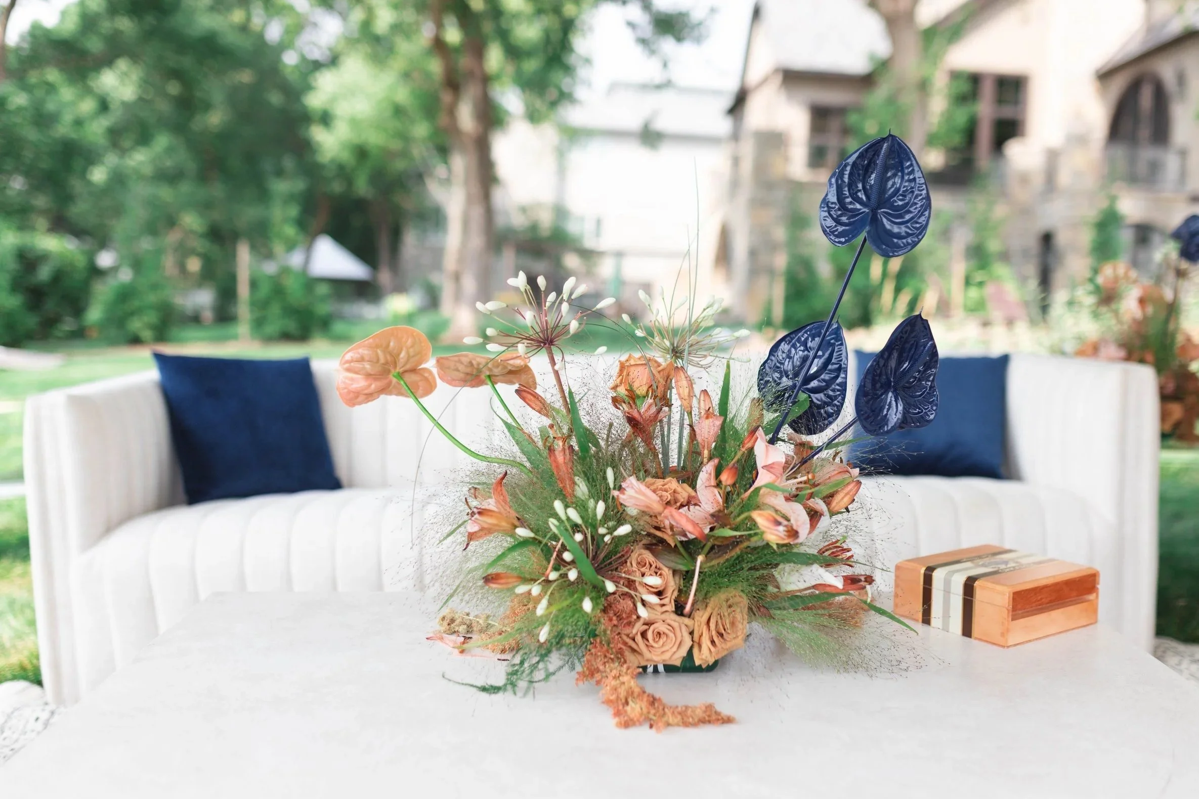 A floral arrangement on a white table in an outdoor patio setting with green trees and a stone building in the background.