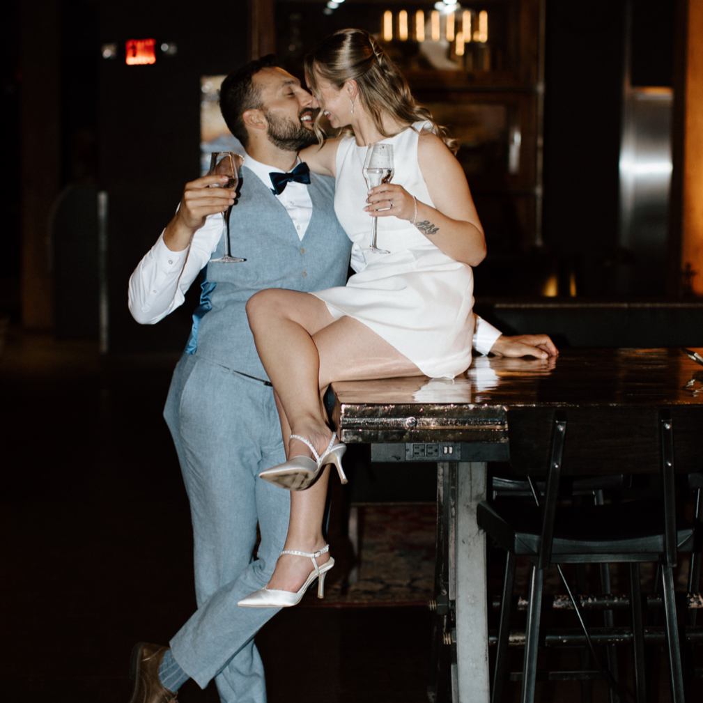 A couple dressed elegantly is celebrating with champagne, sitting on a bar counter in a dimly lit, stylish setting.