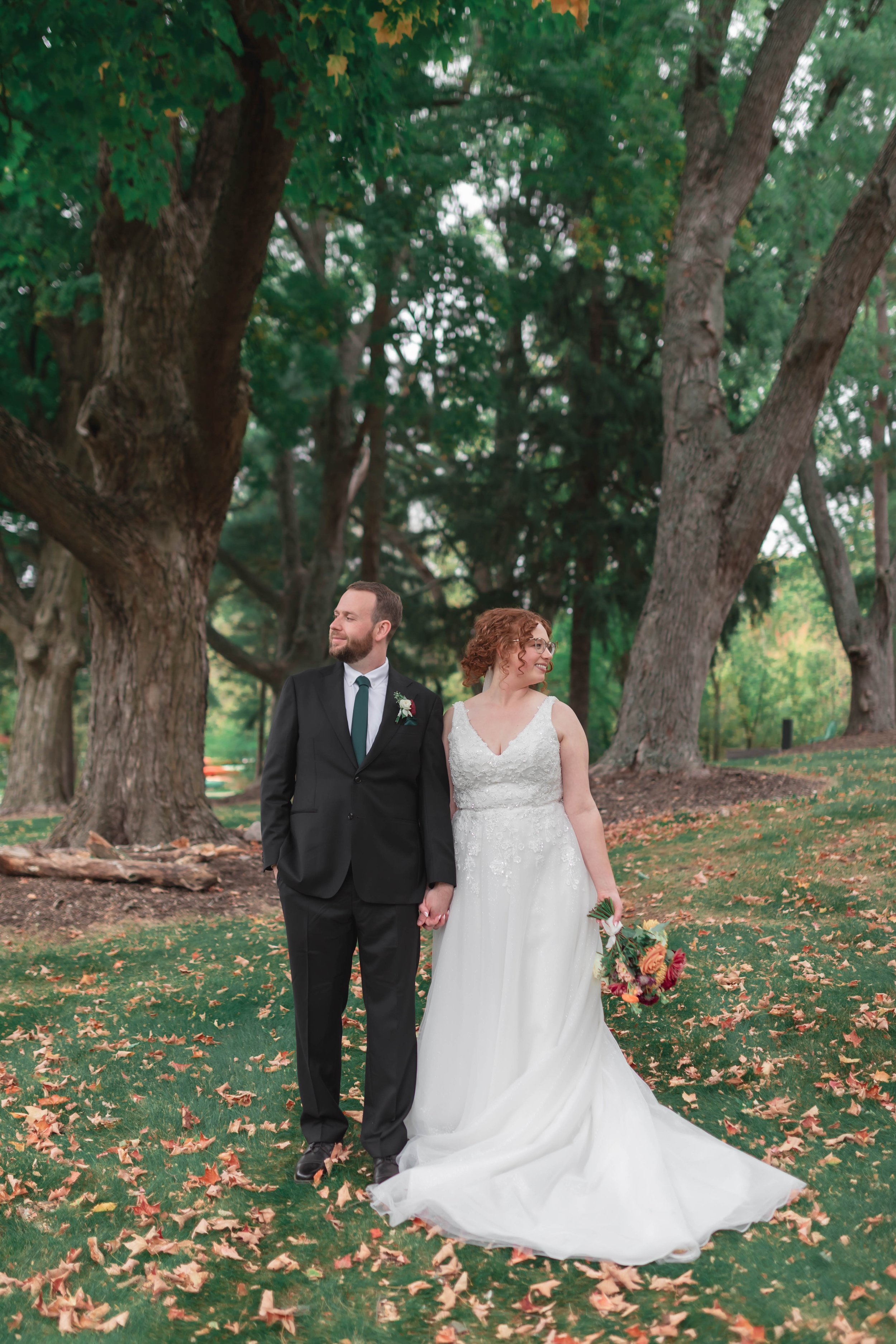 A bride and groom holding hands and walking outdoors in a park surrounded by tall trees with green leaves and fallen autumn leaves on the ground. The groom is wearing a black suit with a white shirt and black tie, and the bride is wearing a white lac