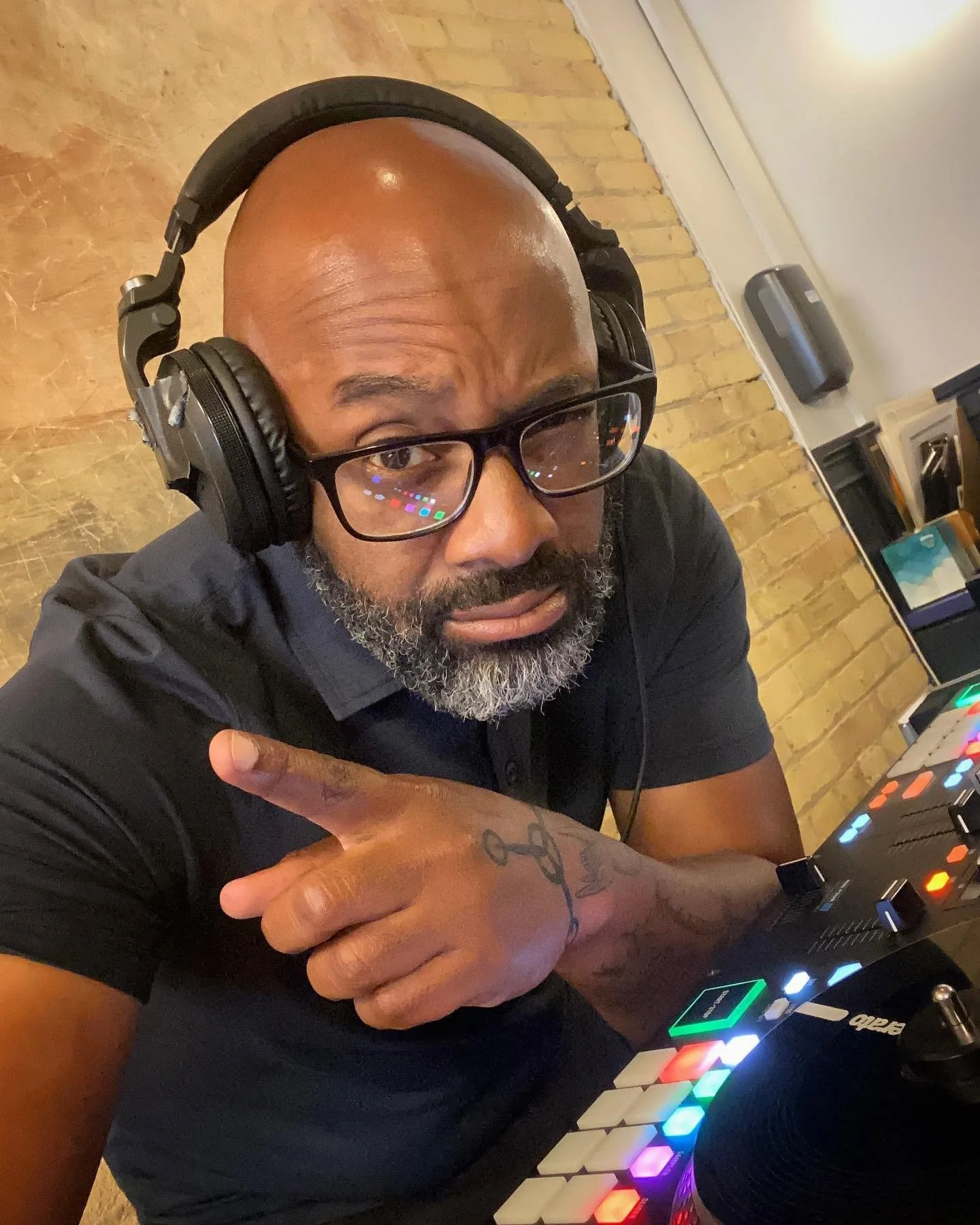 A man with glasses and a beard wearing headphones pointing at a DJ controller with colorful lights.