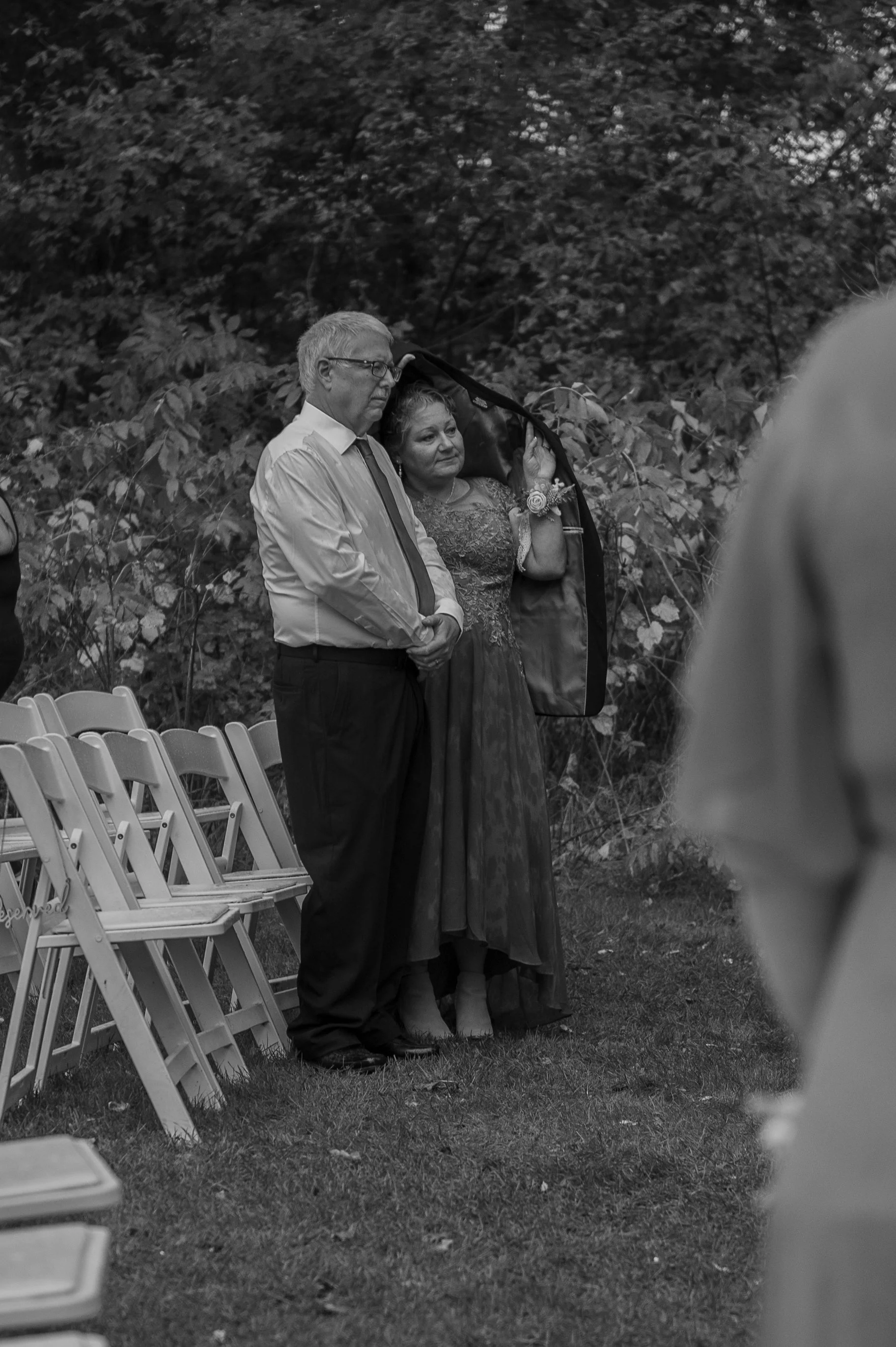 An outdoor wedding ceremony with two people standing in front of rows of empty chairs. The man is wearing a light-colored shirt and dark pants, and the woman is dressed in a lace gown, holding a bouquet and smiling. They are surrounded by trees and f