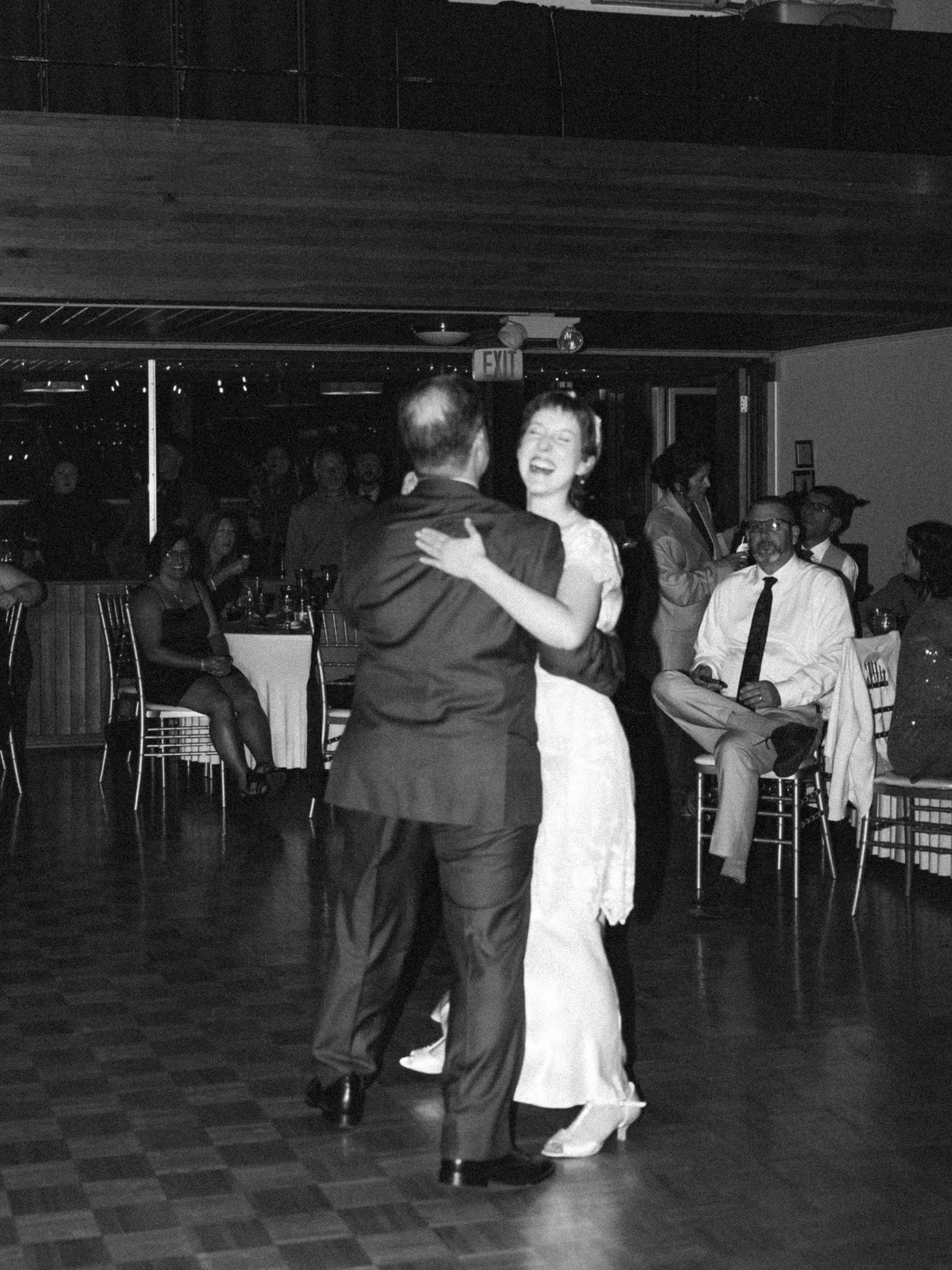 A couple is dancing and laughing at a social event or party, with guests sitting at tables and standing in the background.