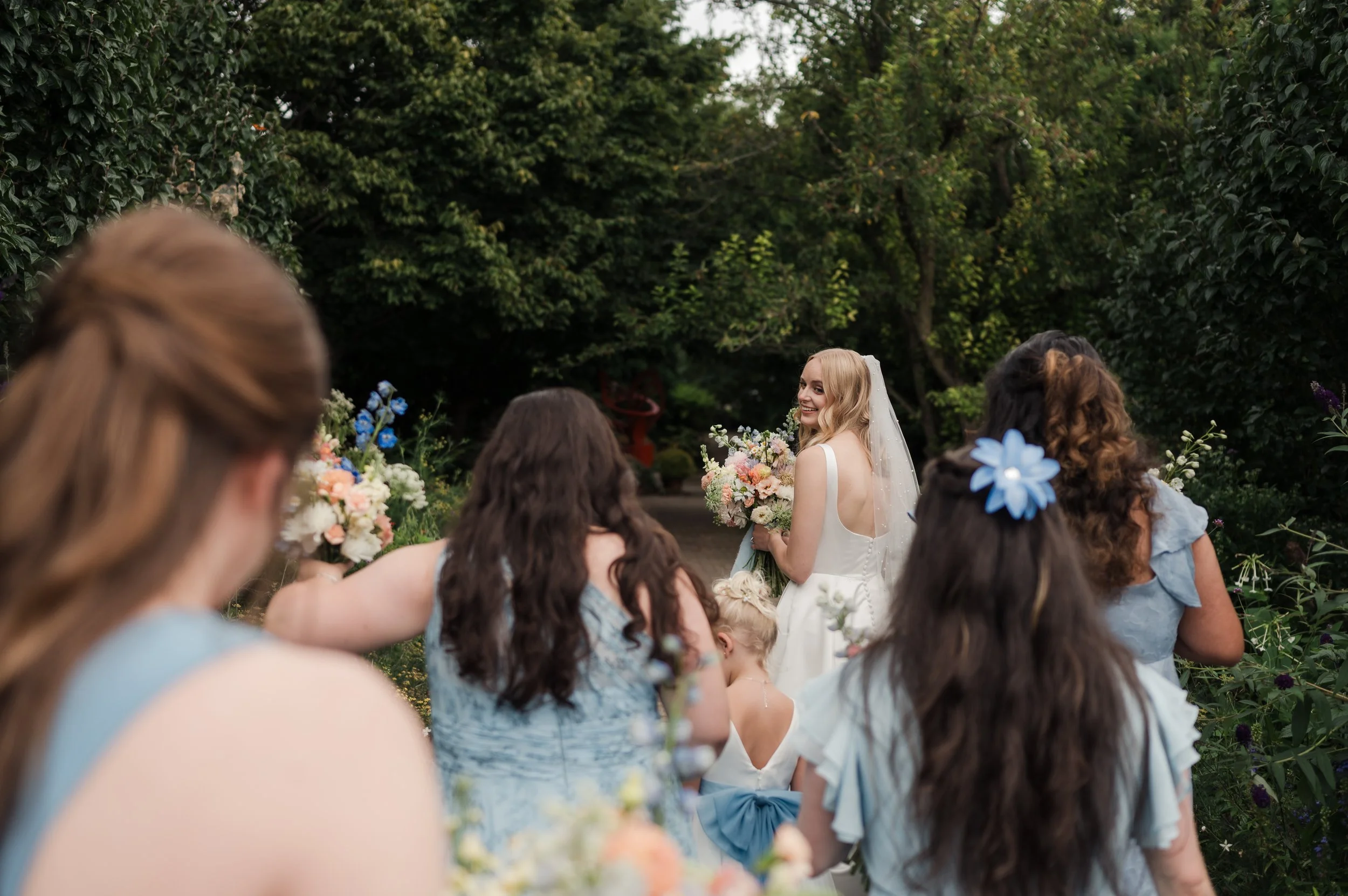 A bride with blond hair holding a bouquet in an outdoor garden surrounded by women in light blue dresses during a wedding celebration.