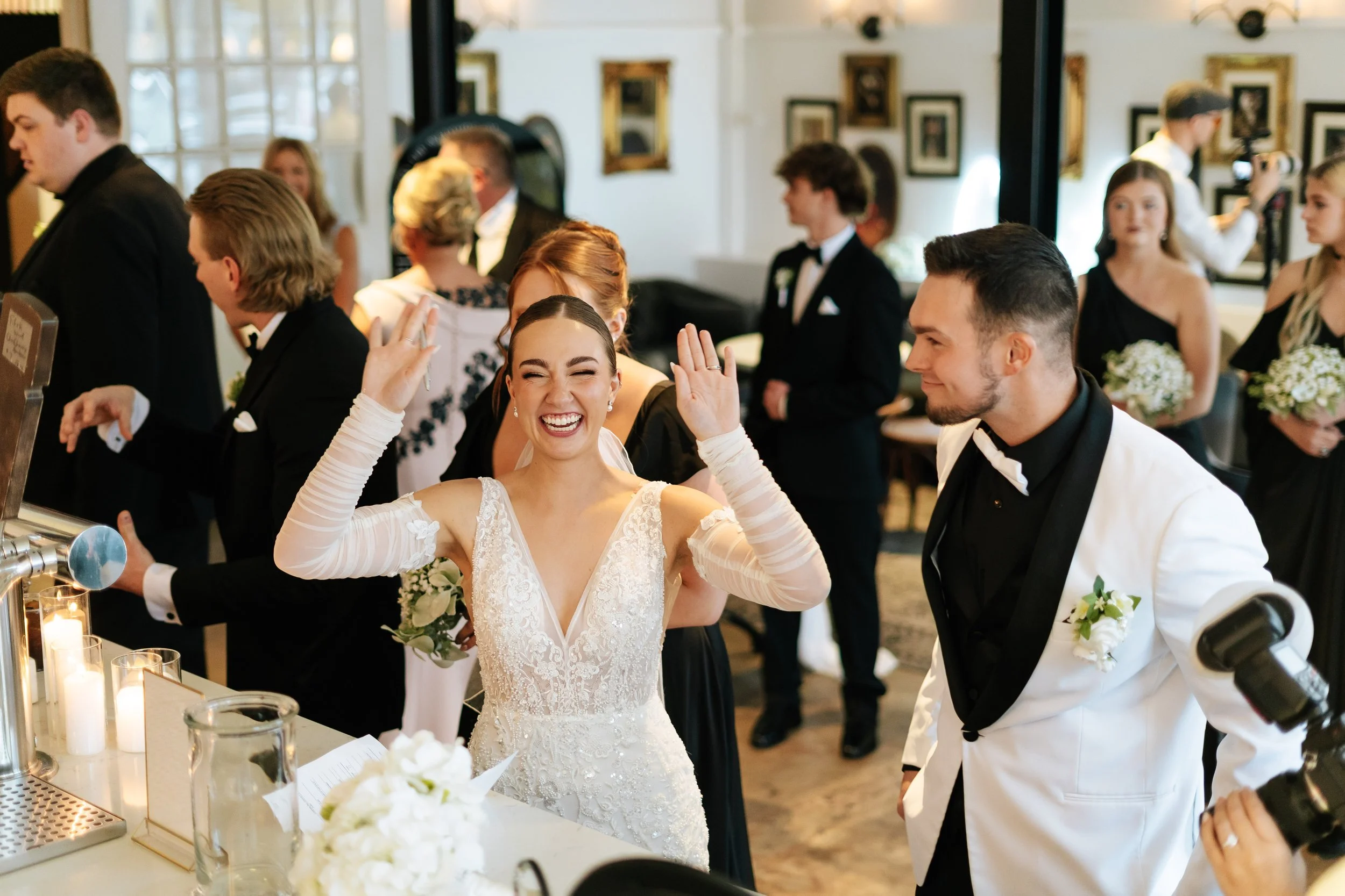 A bride in a white lace wedding gown with long sheer sleeves smiling and raising her hands at a wedding reception, surrounded by guests in formal attire, with some holding bouquets of flowers, in a decorated indoor venue.