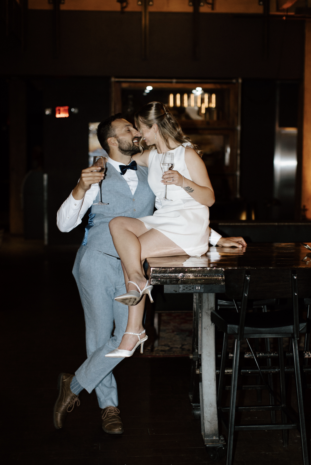 A couple at a celebratory event, with the man wearing a light gray suit and bow tie, and the woman in a white dress, sitting on a table and holding glasses of champagne, sharing a joyful moment