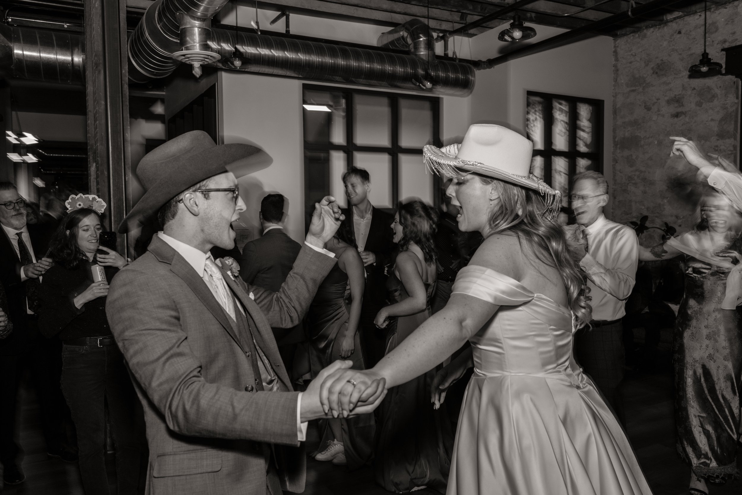 Couple dancing at a celebration with guests in the background, many wearing hats, in an industrial-style venue with exposed ductwork.