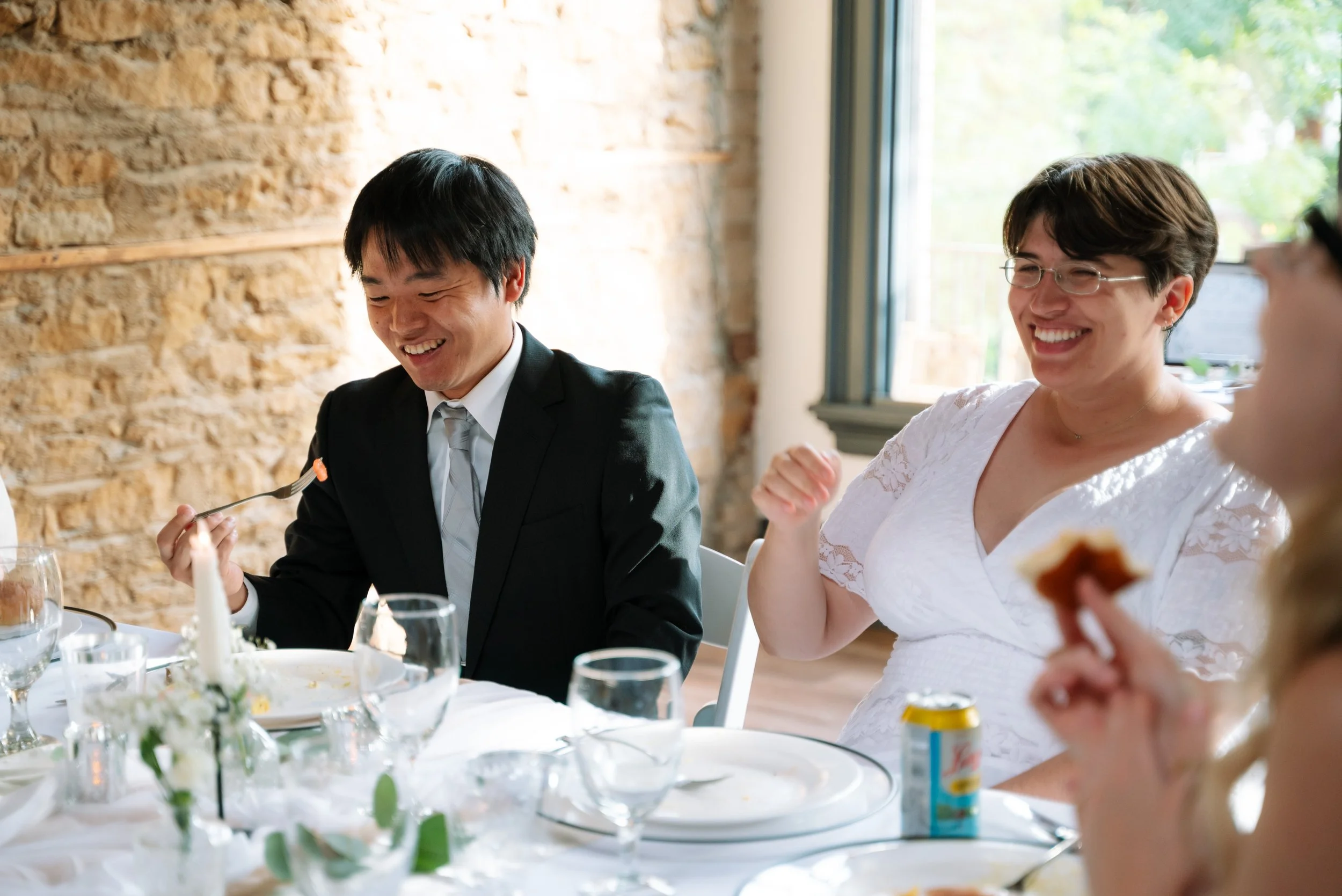 Four people sitting at a dining table, smiling and enjoying a meal together in a well-lit room with a brick wall and large window.