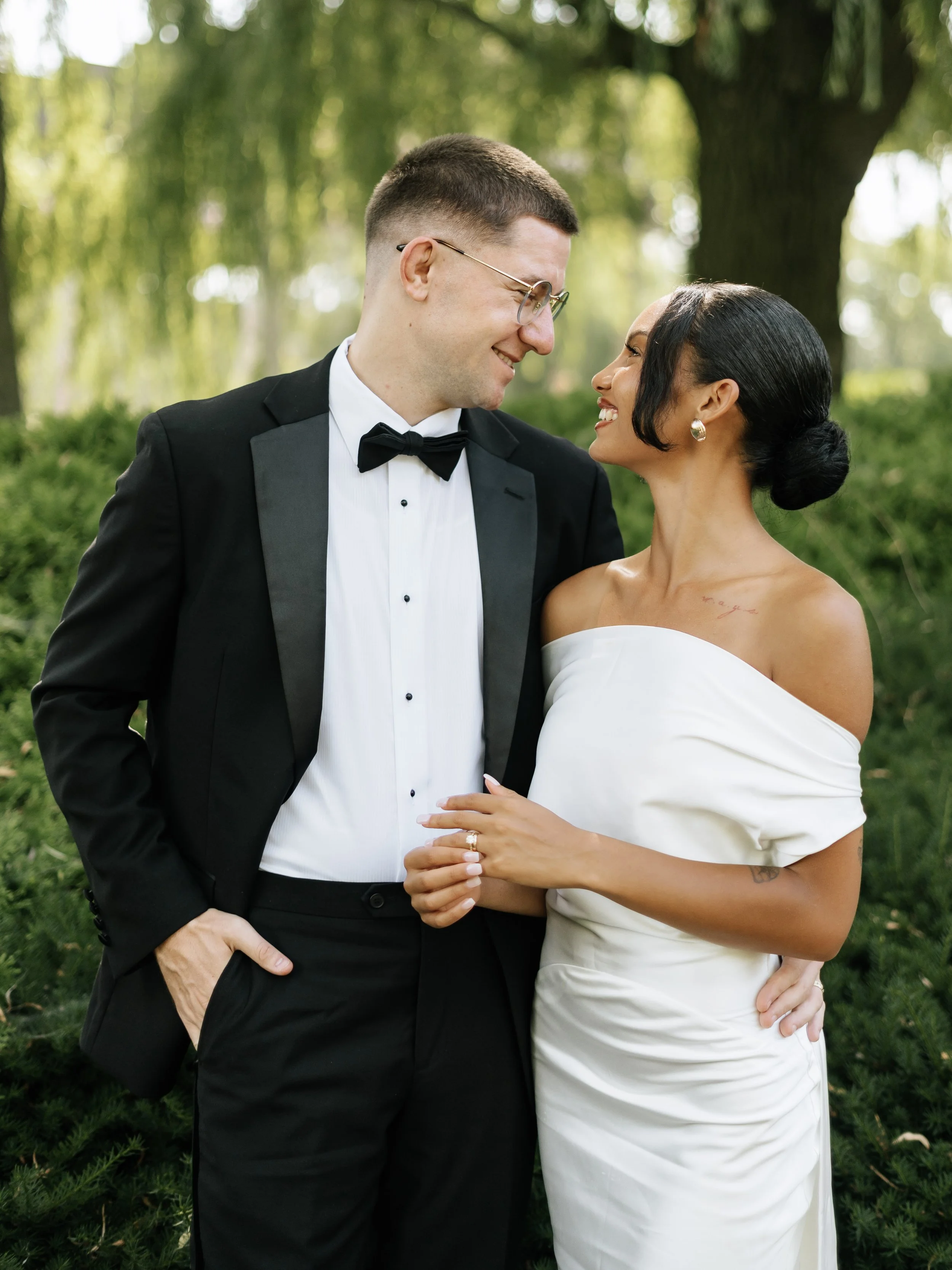A smiling couple in wedding attire, standing close outdoors among greenery, gazing into each other's eyes.