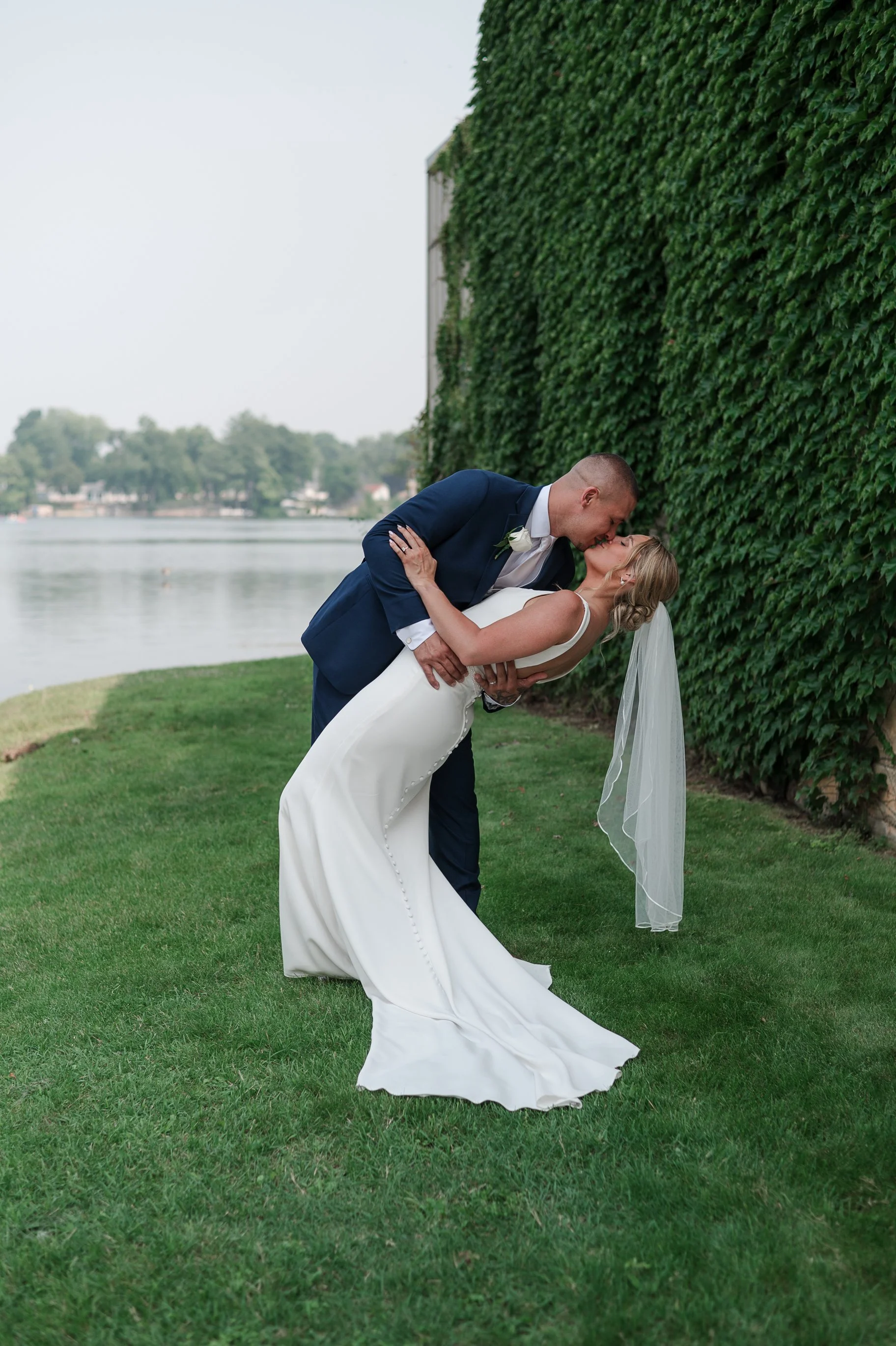 A newlywed couple sharing a kiss outdoors by a river, with the groom dressed in a navy suit and the bride in a white wedding gown and veil, standing on lush green grass next to a tall hedge.