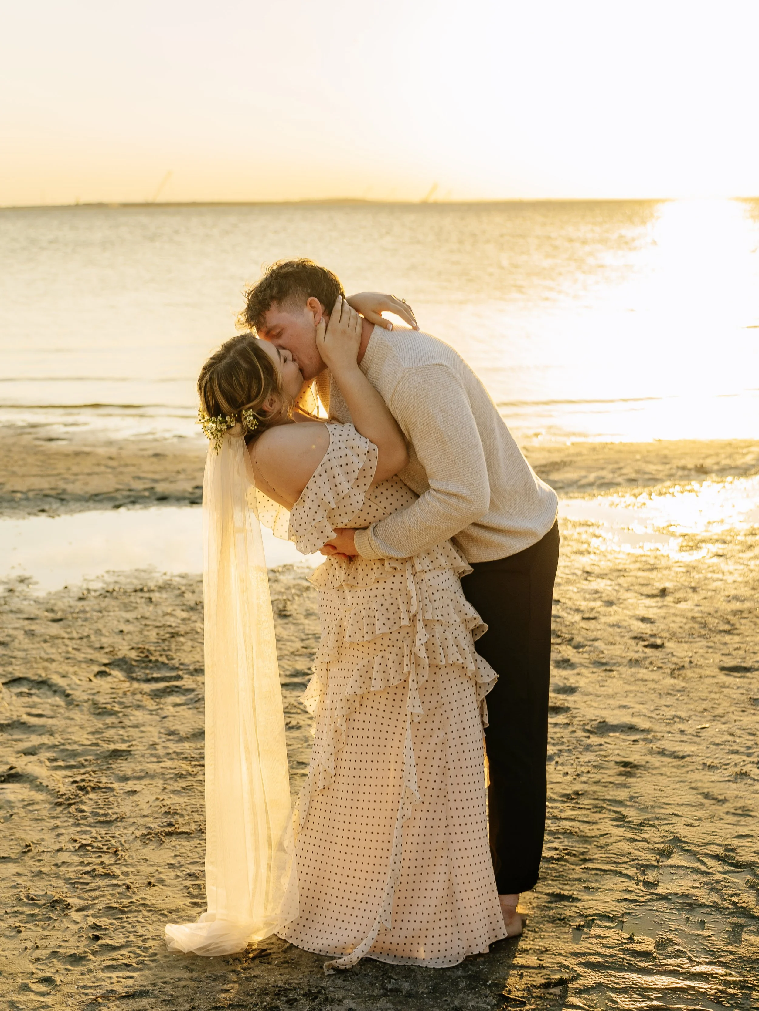 A couple kissing on the beach during sunset, the woman wearing a beige polka dot dress and a veil, the man in a light sweater and dark pants.