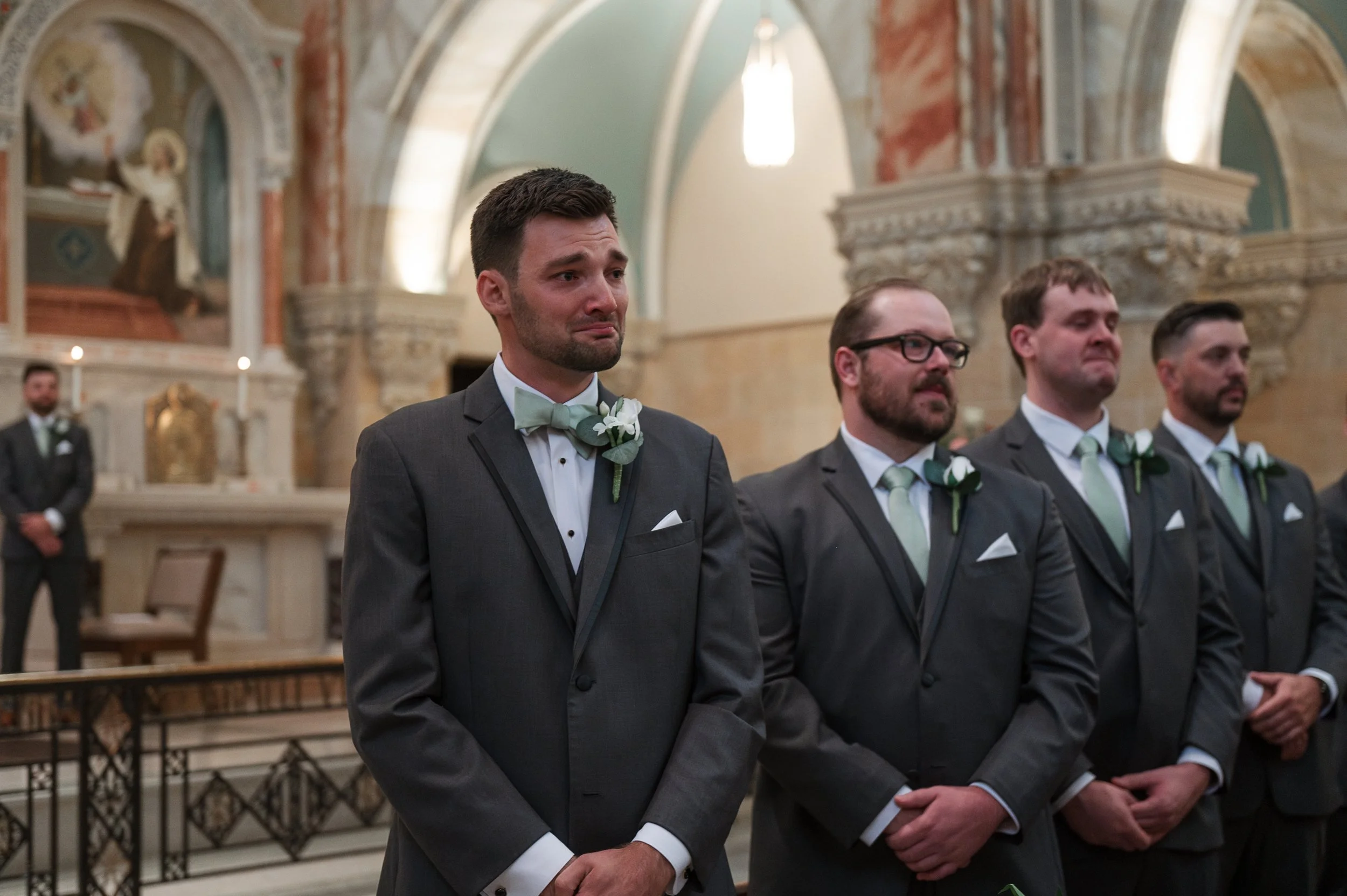 A group of men in tuxedos with green ties and white boutonnières standing at a wedding ceremony in a church, with one man appearing emotional, possibly crying.