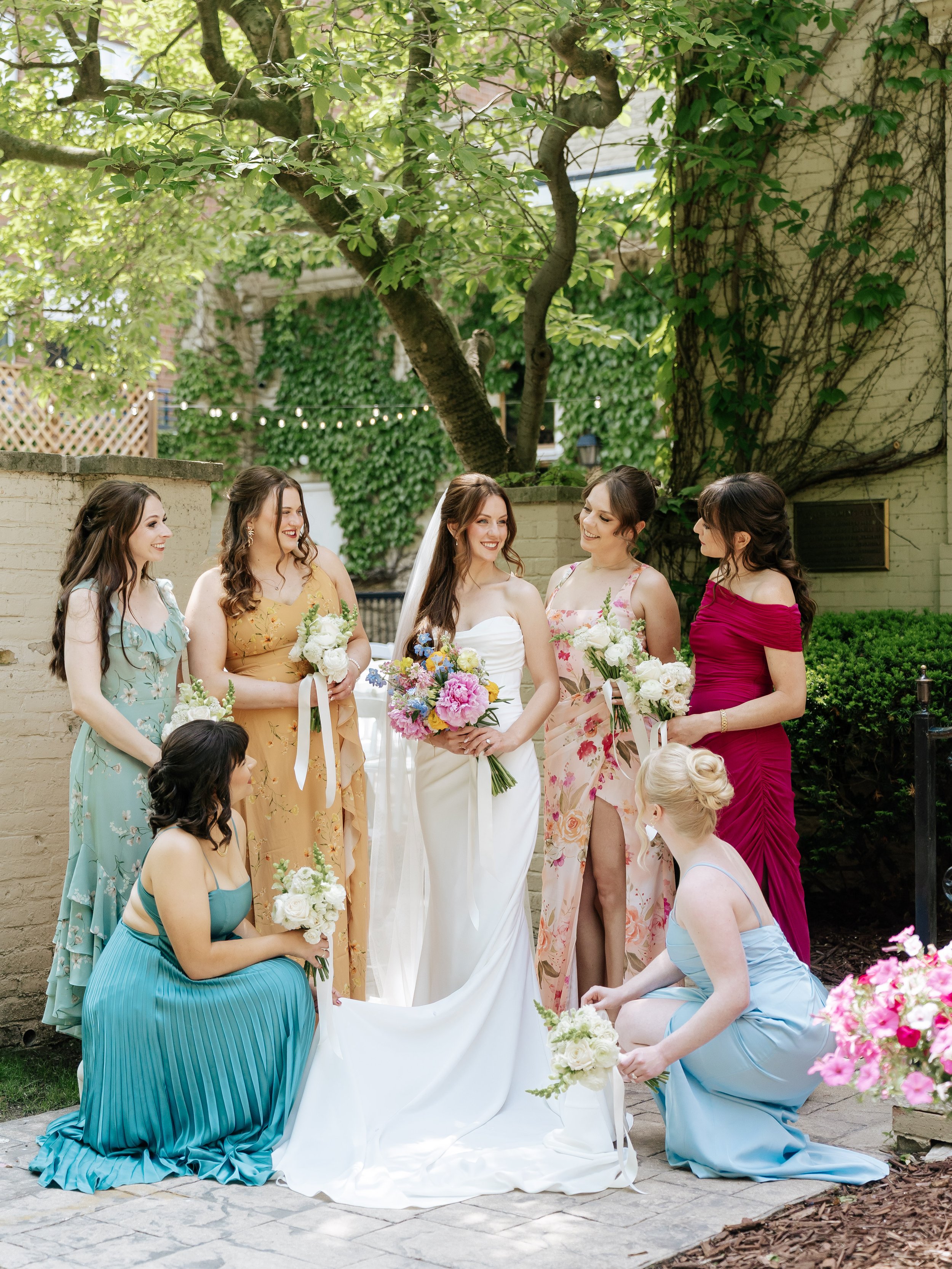 A group of women at a wedding, with the bride in white at the center holding a bouquet and surrounded by bridesmaids dressed in colorful gowns holding bouquets, outdoors with lush greenery and a tree in the background.