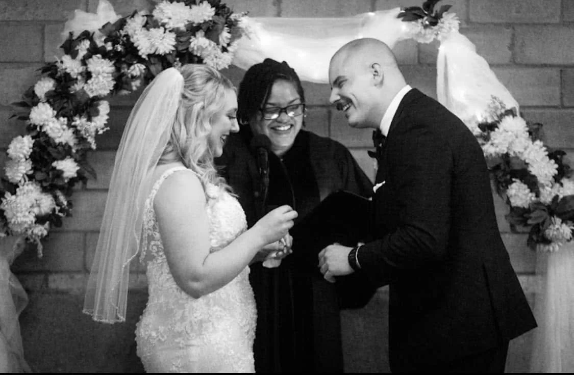 Bride and groom exchange vows under a floral arch, smiling at each other, during their wedding ceremony with officiant presiding.