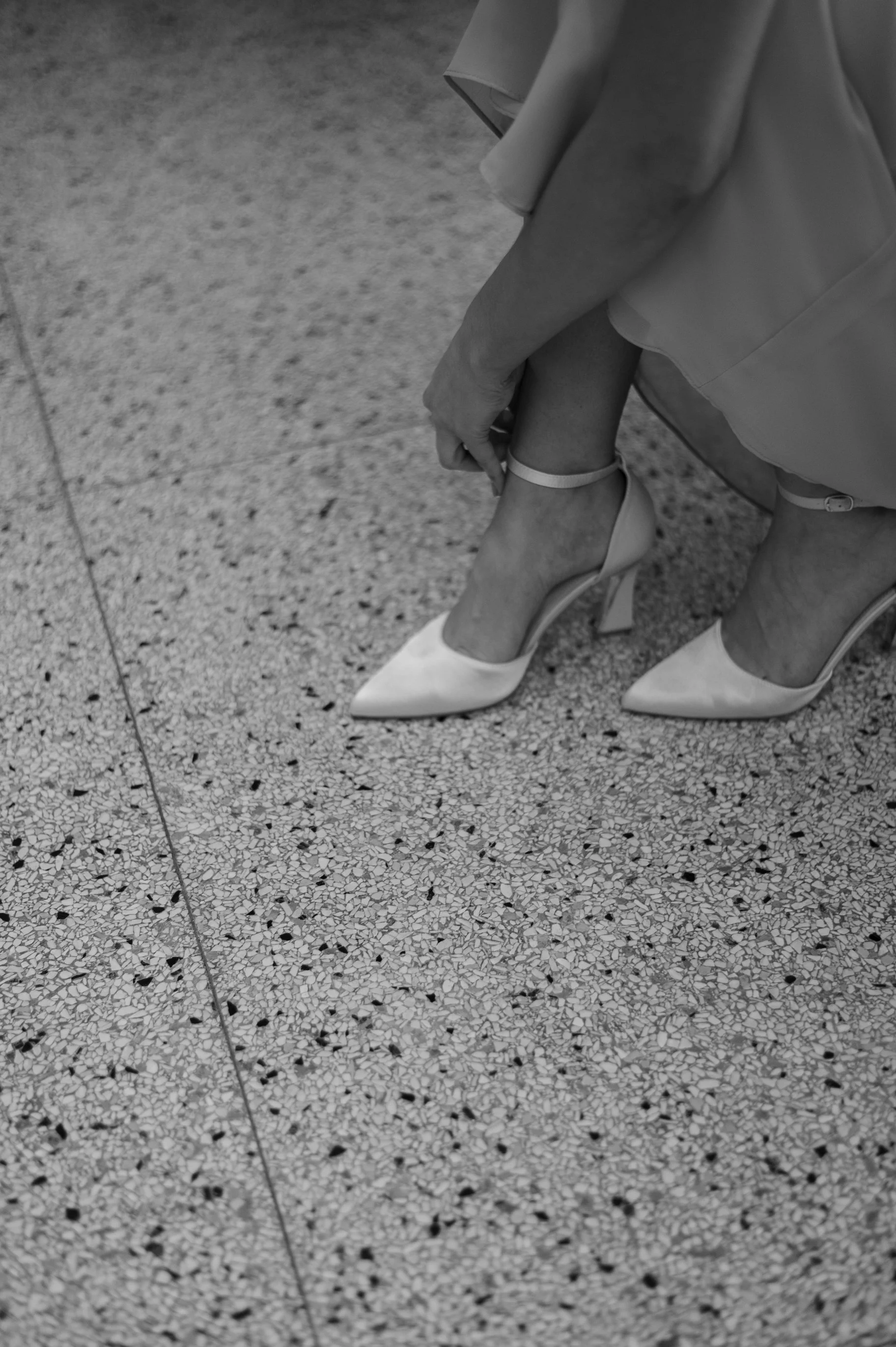 A person adjusting their ankle strap on a high-heeled shoe while sitting on a terrazzo floor.