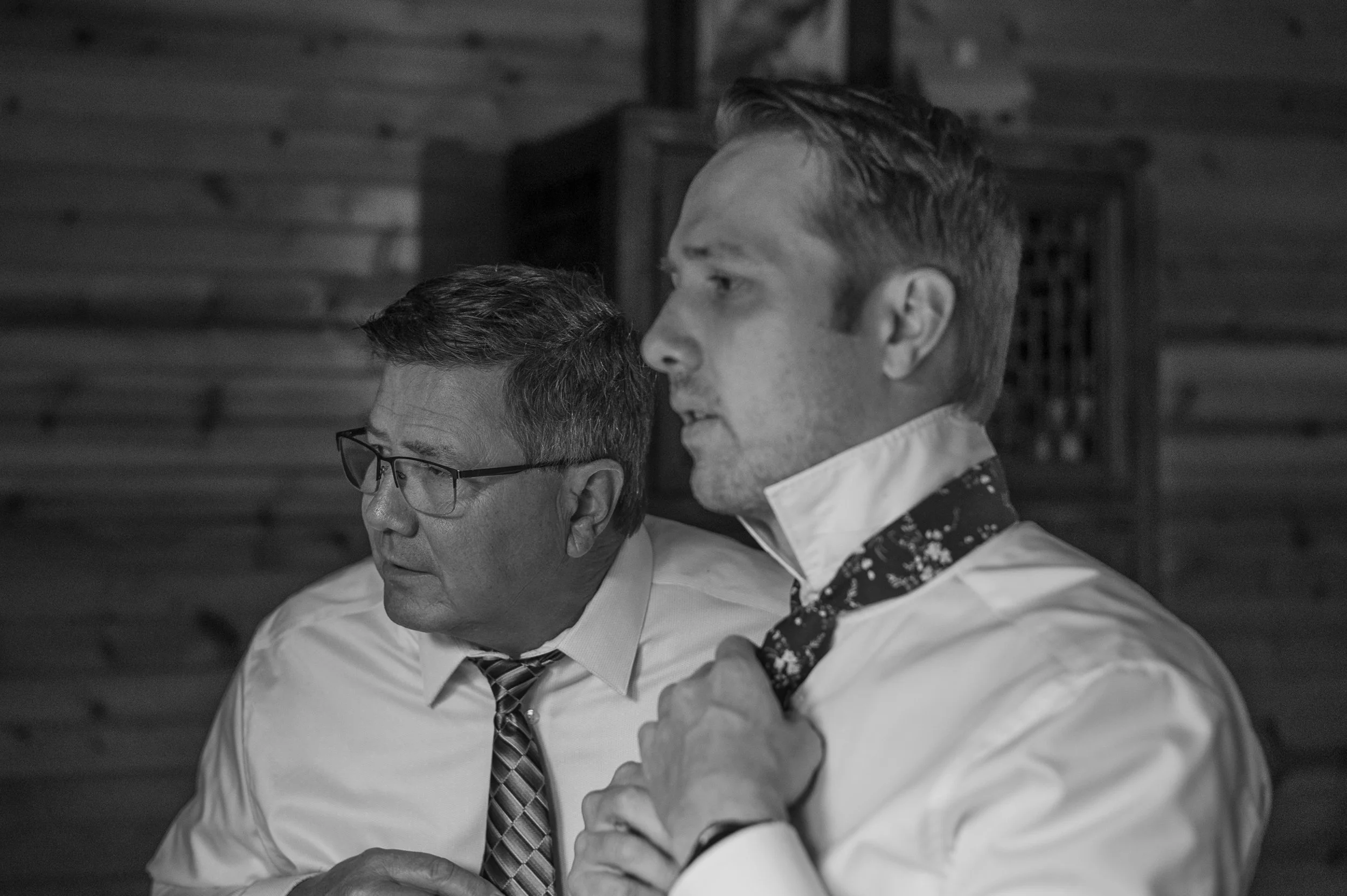 Two men dressed in formal attire, one with glasses and the other adjusting his floral patterned tie, in a room with wooden walls.
