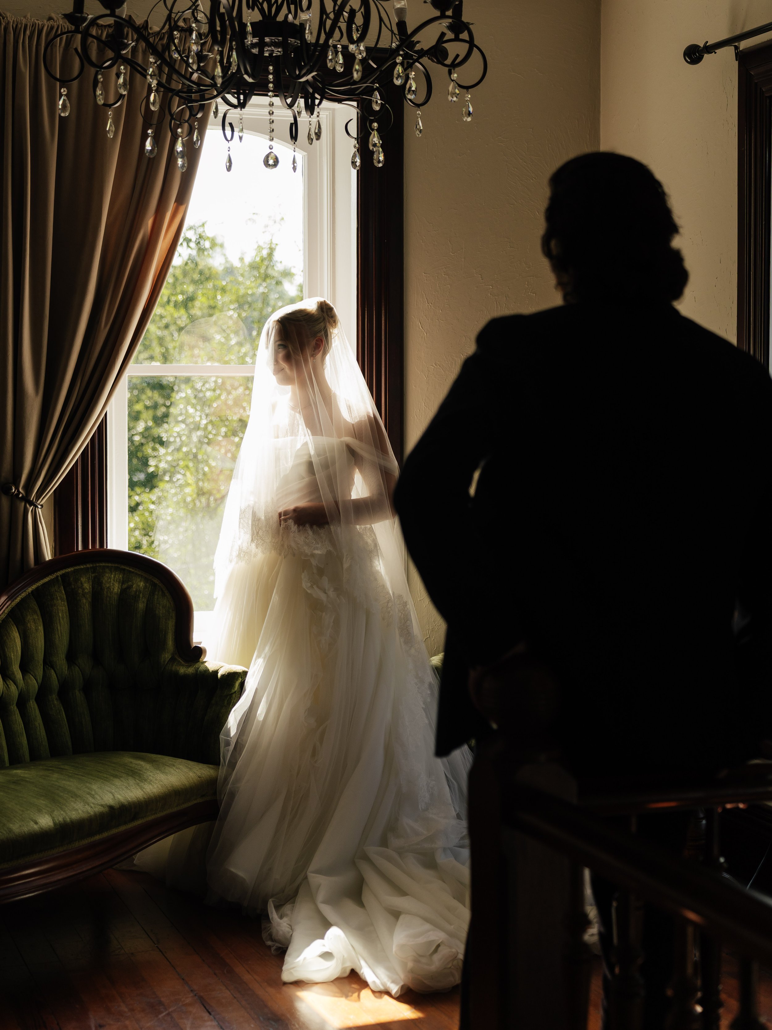 Silhouette of a man and a bride in a wedding dress, with the bride standing near a window with sunlight shining through, highlighting her veil and gown.