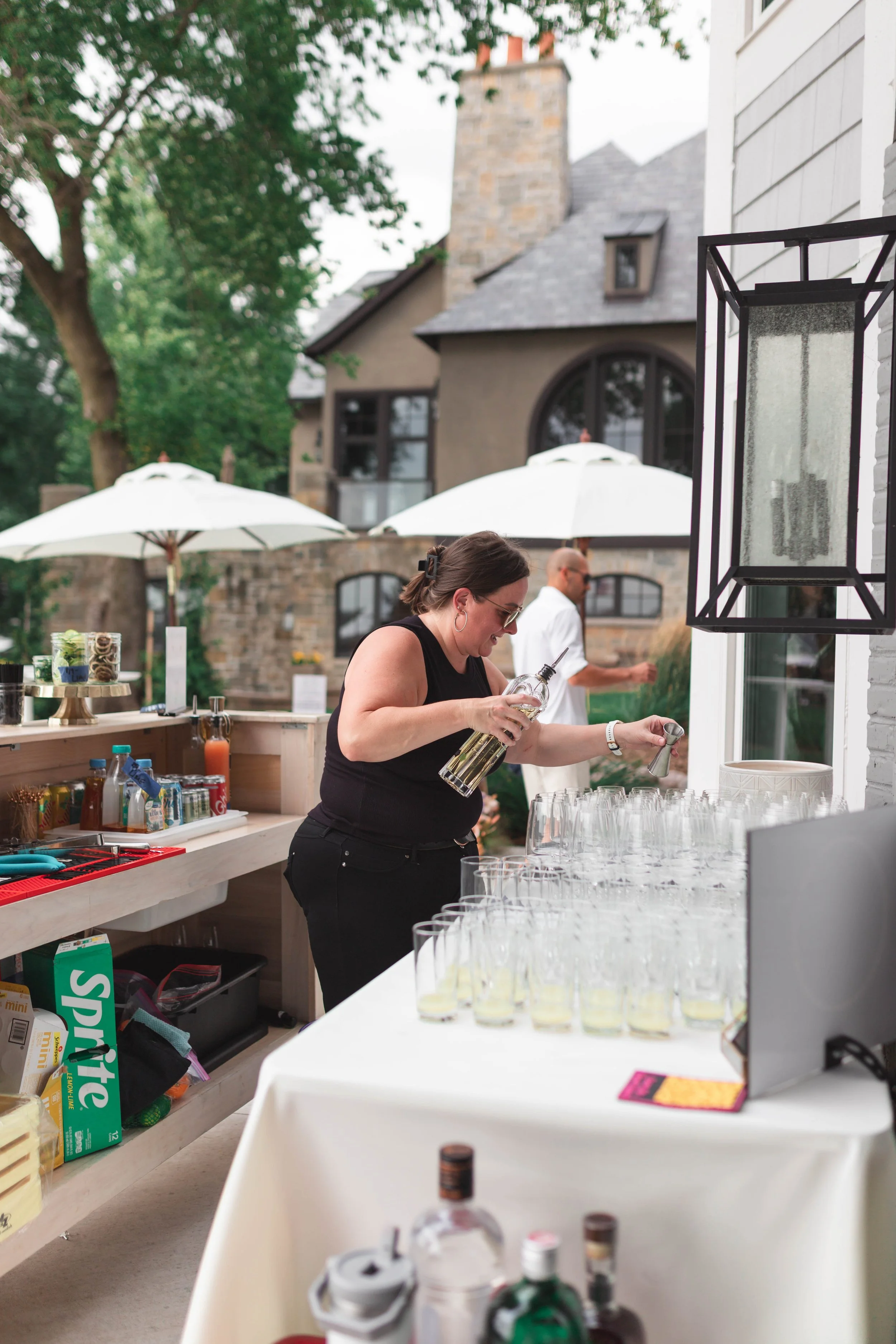 A woman in a black top pouring a drink into a glass at an outdoor setup with white umbrellas and a stone house in the background.