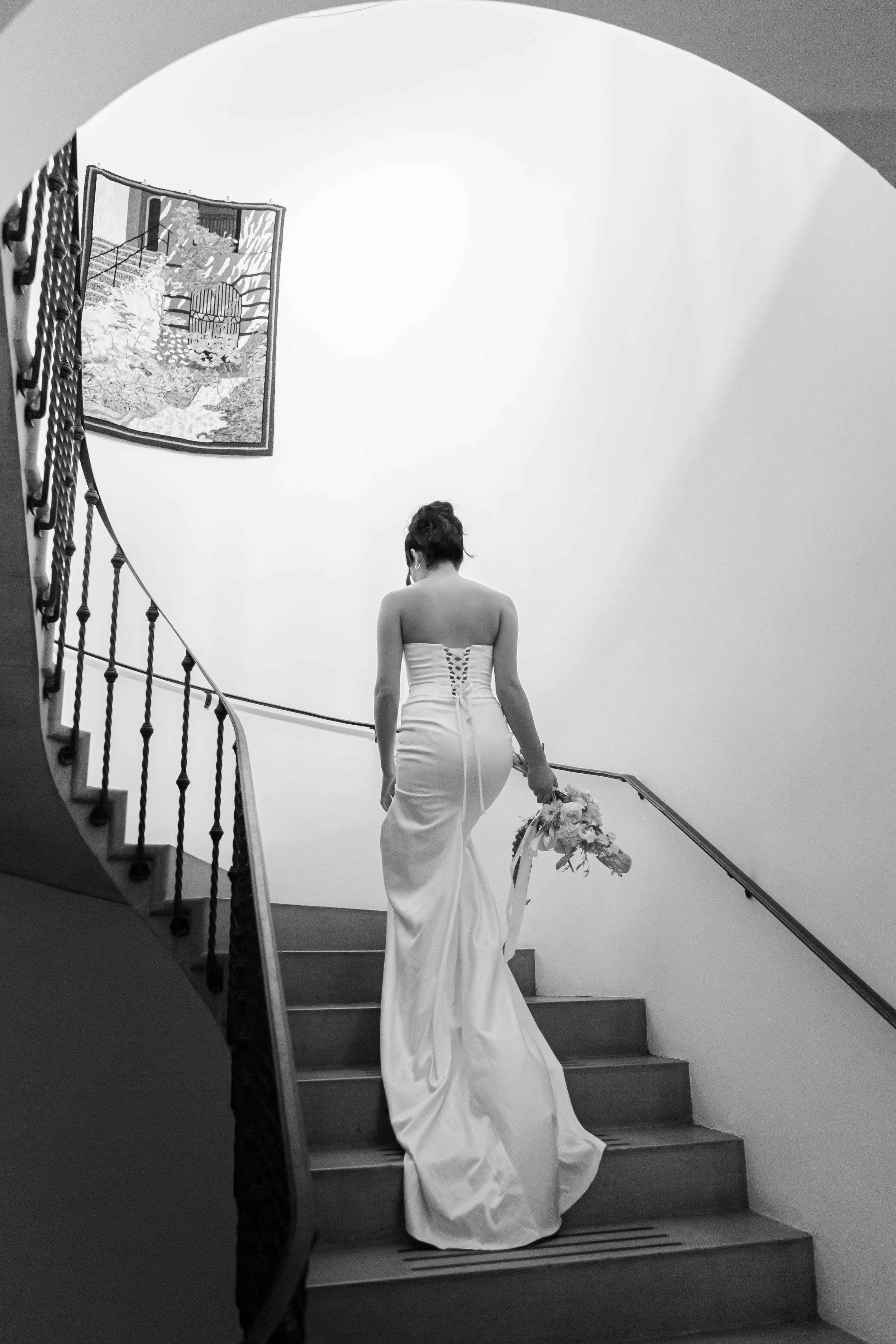 A woman in a wedding dress walking down a staircase while holding a bouquet, viewed from the back in black and white.