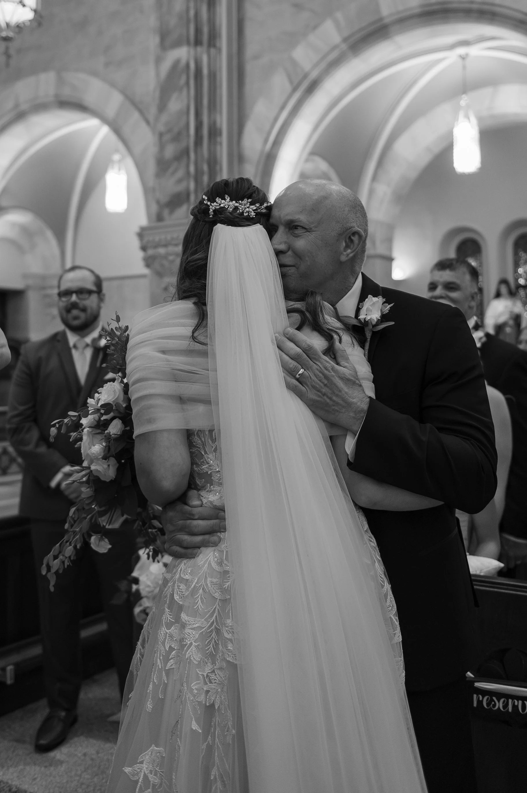 A bride and a man, likely her father, sharing an emotional hug during a wedding ceremony in a church, with other guests smiling in the background.